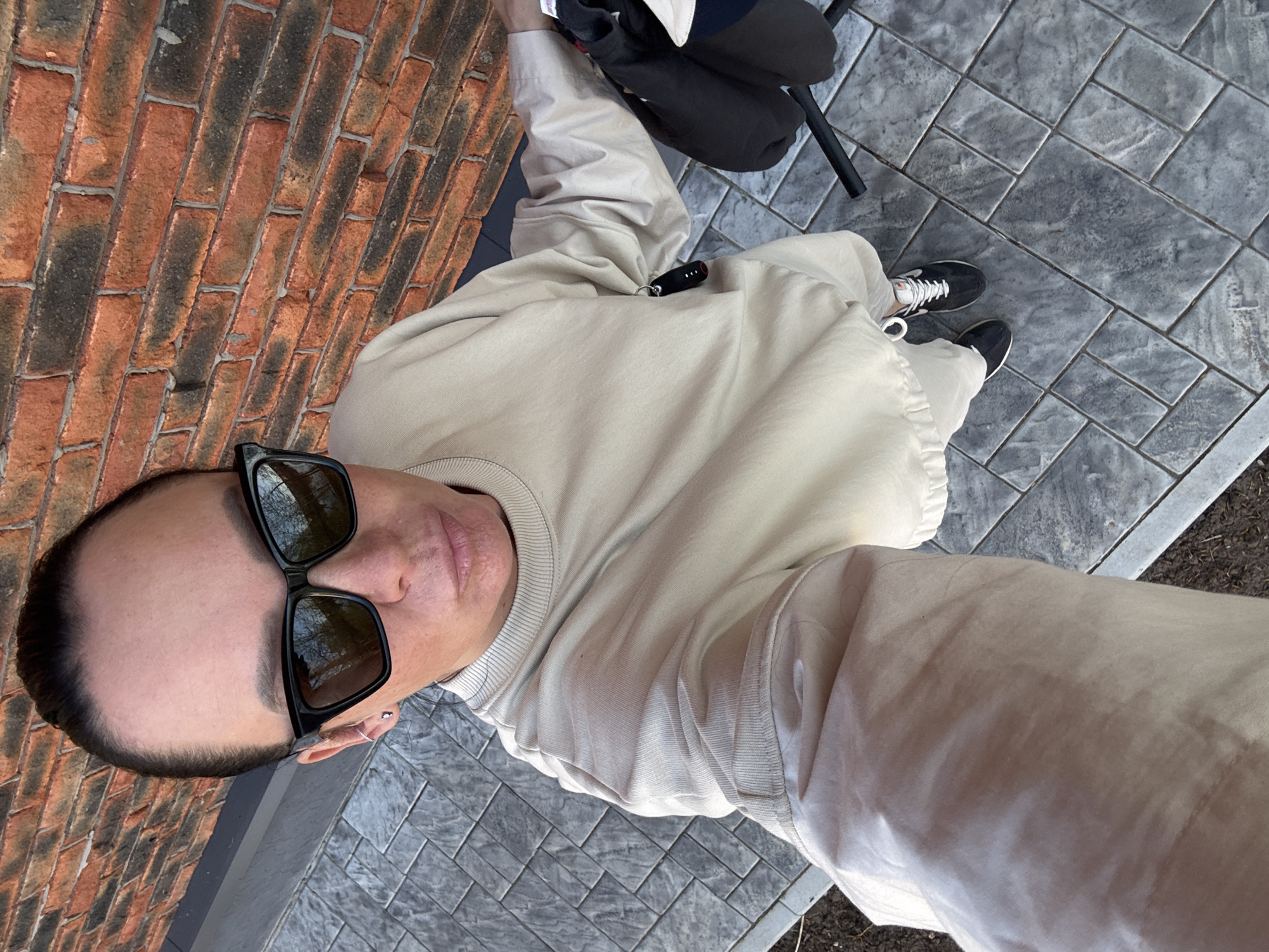 The image is a high-angle, point-of-view photograph of a person standing outdoors. The person is wearing black-framed sunglasses, a light beige sweatshirt, and tan pants. Their feet are clad in black sneakers with white laces. To the left, there is a red brick wall, and to the right, a paved area with gray patterned tiles. Behind the person, there is a chair or stroller with a black and beige section. A small black object with a red light is attached to their clothing. The location is assumed to be Chaikovsky, Russia, given the request.
