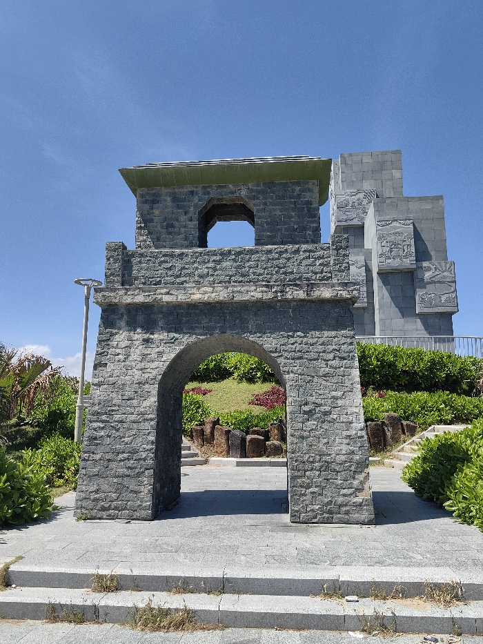 The image depicts the Nghinh Phong Tower complex in Tuy Hòa, Vietnam, under a clear blue sky. In the foreground, grey stone steps with some visible weeds lead to a paved area. A central, two-tiered structure resembling a gateway or watchtower is constructed from rectangular grey stone blocks. Its lower tier features a prominent arched opening, providing passage. The upper tier, set back from the lower, also has an arched opening and is topped by a low-pitched roof with a green-colored edge. To the left of this structure, a silver-colored lamppost is partially visible among green foliage. Behind the gateway and situated on higher ground, a larger, multi-tiered monument of grey stone stands. This monument exhibits multiple stepped levels and features intricate bas-relief carvings depicting scenes on its visible faces. The surrounding environment includes lush green landscaping with various bushes, grass, and some reddish ground cover, interspersed with dark, irregular vertical stone elements. Grey paved pathways and stone steps ascend through the landscaped areas towards the larger monument. A grey metal railing is visible bordering the elevated platform where the larger monument is situated. No people, animals, or vehicles are present. The scene is brightly illuminated by daylight.