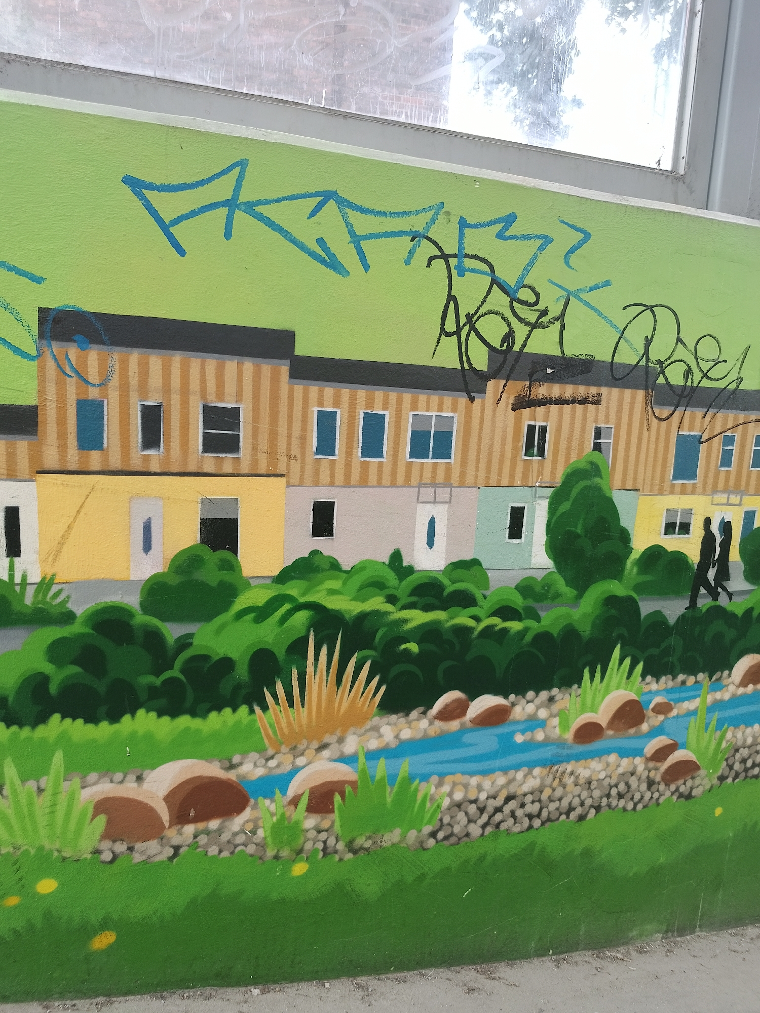 The image shows a mural depicting a row of houses along a waterway. The houses are painted in alternating colors of yellow, light gray, and light blue, with brown striped walls and black roofs. Each house has windows and doors. The waterway is depicted with blue water, small gray stones lining the banks, and brown rocks scattered throughout. Green vegetation, including bushes, tall grasses, and other greenery, lines the waterway and the front of the houses. Two figures in silhouette walk along the pathway in front of the houses. Above the houses and trees, graffiti in blue and black is visible against a light green background. A window frame is visible above the mural. The city and precise location are unknown.
