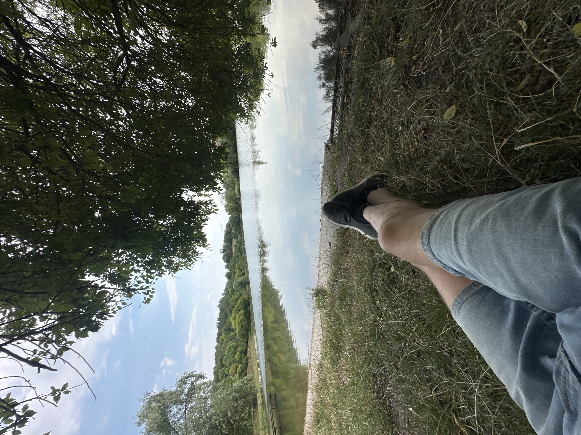 The image depicts a serene lakeside scene on a sunny day, likely during summer in Makiv, Ukraine. In the foreground, a person's legs are casually crossed, clad in denim shorts and black sneakers, resting on dry grass. The person is seated under the shade of lush green trees whose branches extend into the frame, obscuring some of the sky. The middle ground features a tranquil lake with a calm surface reflecting the blue sky and scattered white clouds. The shoreline is visible, with a sandy or gravelly edge leading to the water, and some vegetation. A wooden structure, possibly a pier or a jetty, is partially submerged in the water on the left side of the lake. The background is a picturesque landscape of rolling hills covered in dense green forests. The overall atmosphere is one of relaxation and enjoyment of nature.