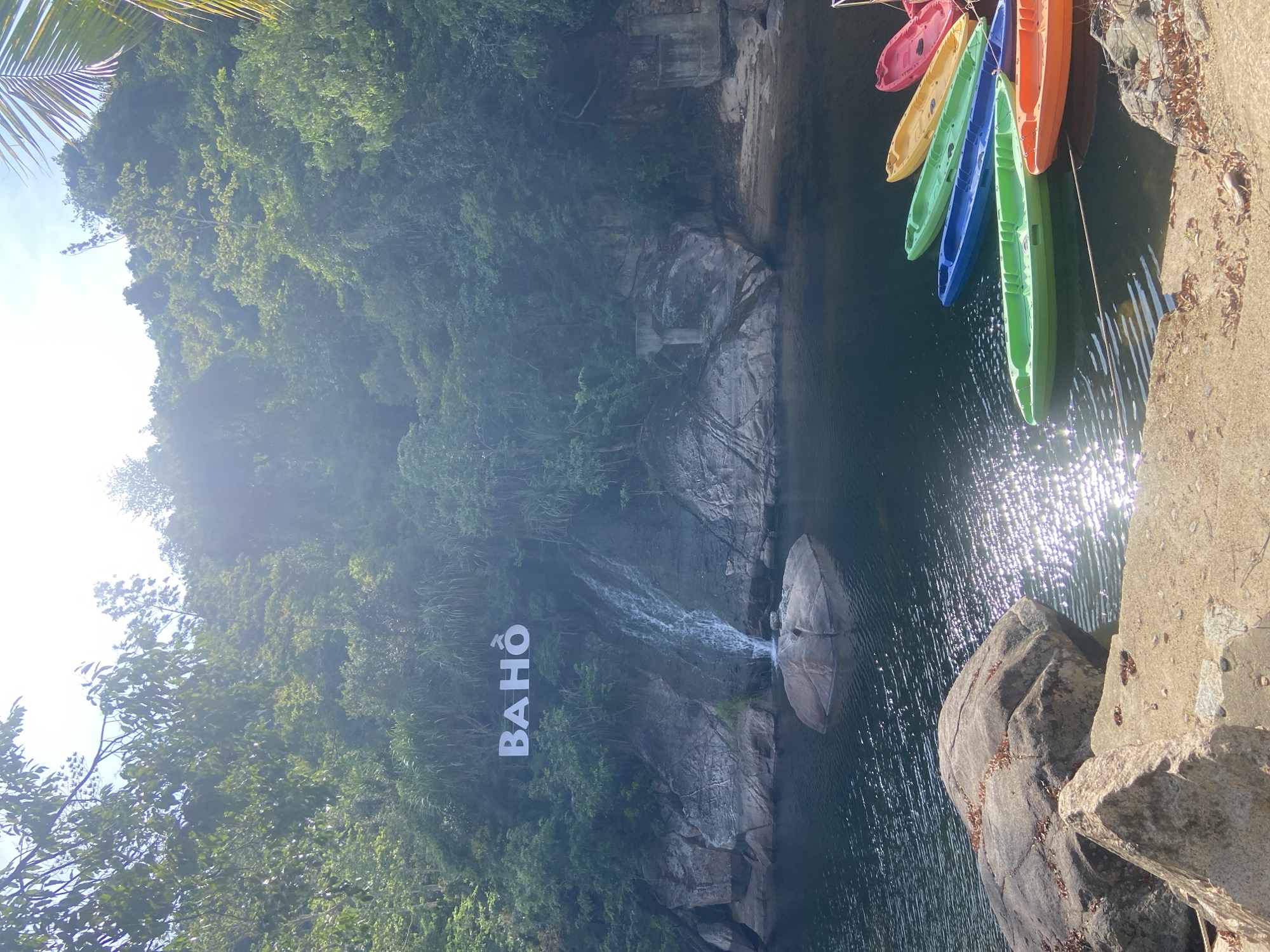 The image depicts a scenic view of the Ba Ho waterfalls located in Hoa Thang Ward, Vietnam. In the foreground, several brightly colored kayaks are moored on the dark water of a river, including orange, blue, green, yellow, and pink vessels. The river's surface is illuminated by shimmering sunlight. On the left side of the frame, large rocks line the shore, with one prominent boulder in the water near the base of the waterfall. The waterfall itself cascades down a rocky cliff face, with the word "BA HO" displayed in large white letters on the hillside above it. The surrounding environment is lush and verdant, with dense green foliage and trees covering the slopes of the hills. The sky above is bright and partly obscured by sunlight and foliage.