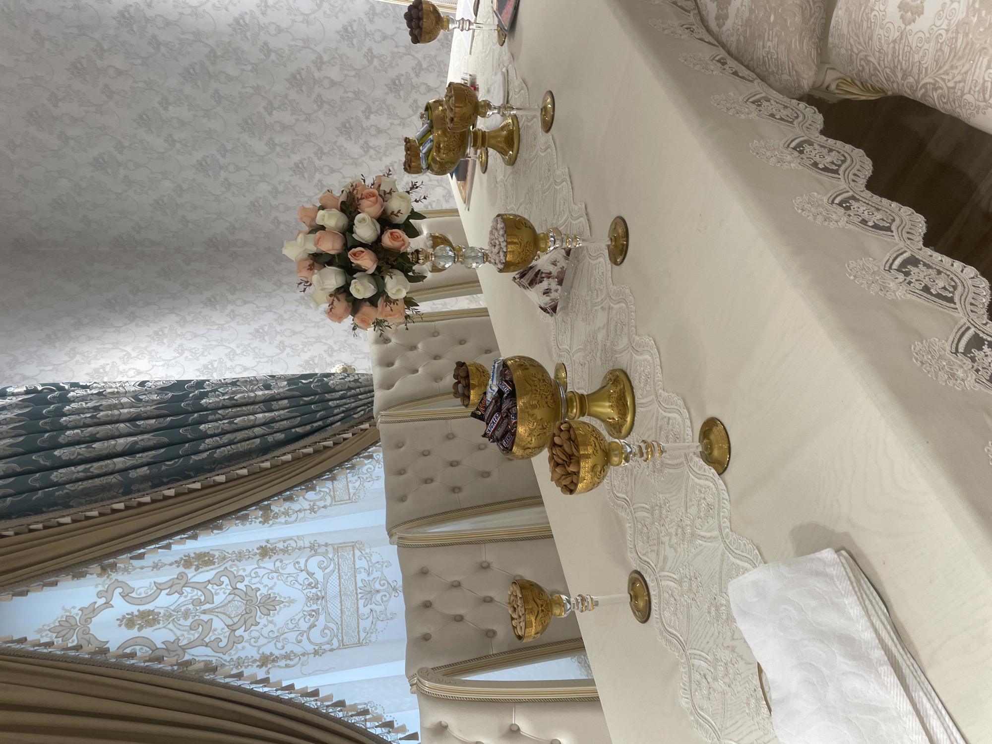 The image depicts a formal dining table set for an occasion, likely a celebration or gathering, in Tashkent, Uzbekistan. The table is covered with a light-colored tablecloth adorned with an intricate lace runner. Several ornate gold bowls, some on pedestals, are strategically placed on the table, filled with assorted nuts and chocolates. One prominent gold bowl contains an assortment of chocolate bars. A bouquet of artificial roses in shades of pink and white adds a touch of color to the scene.
The table is surrounded by elegant, tufted upholstered chairs with gold trim. In the background, a window is dressed with layered curtains: sheer white patterned curtains are behind heavy gold drapes, and a darker teal curtain is visible on the right side. The wall behind the table features textured wallpaper with a damask pattern. The overall impression is one of refined opulence and careful presentation. No people are visible in the image. The lighting suggests an indoor setting, possibly during daylight hours given the illumination from the window. The quality of the image is high, with good detail and color rendition.