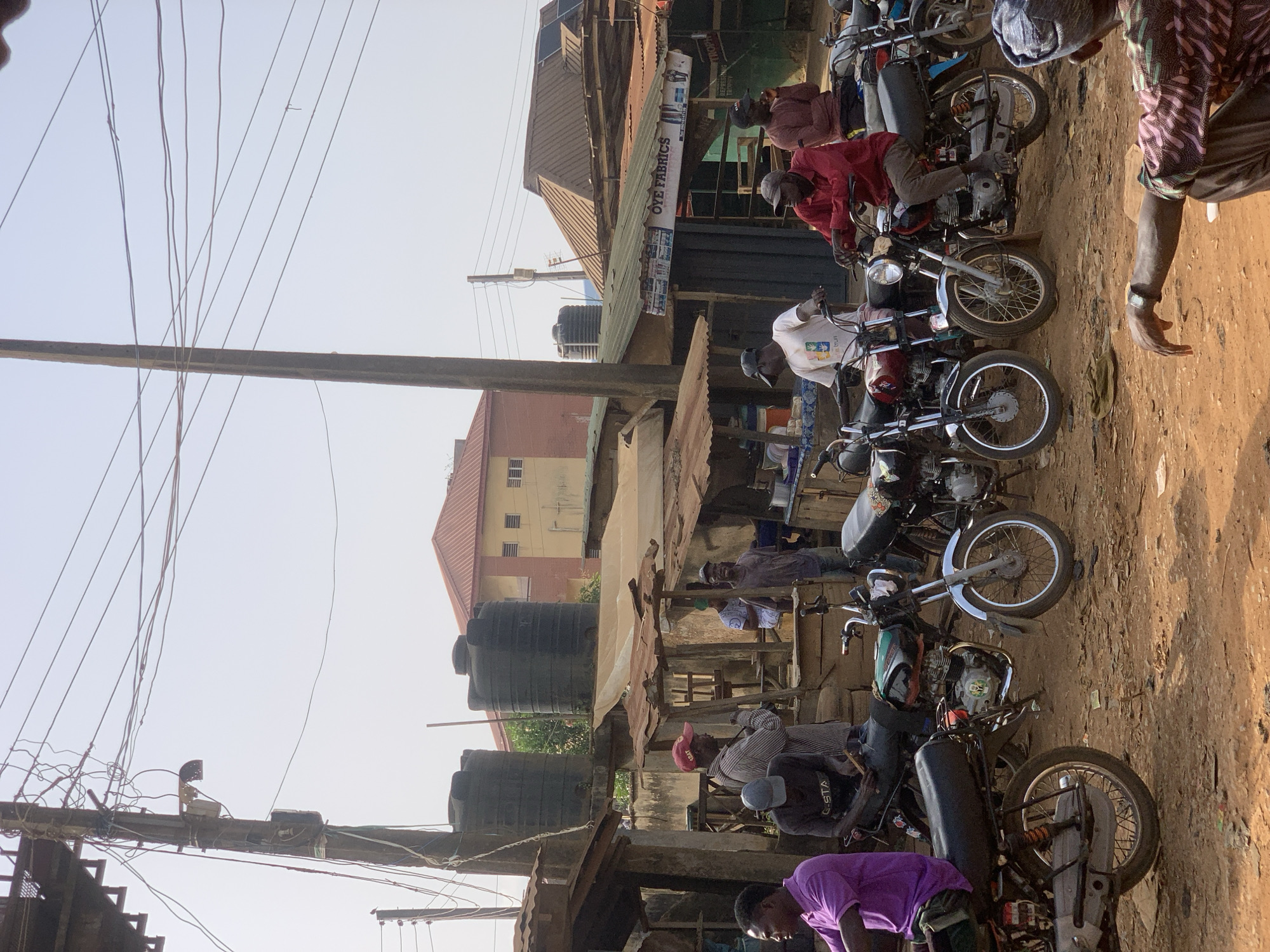 The image captures a street scene in Ikorodu, Nigeria. A row of motorcycles, also known as Okada, are parked along a dirt road. Riders or passengers are seated on some of the motorcycles, some wearing casual clothing. The buildings on the left of the image are older structures with corrugated iron roofs and large black water tanks. Electrical wires crisscross the sky. A business sign with "OYE FABRICS" is visible. A partially visible red building is in the background on the left. The ground is dirt and dust.