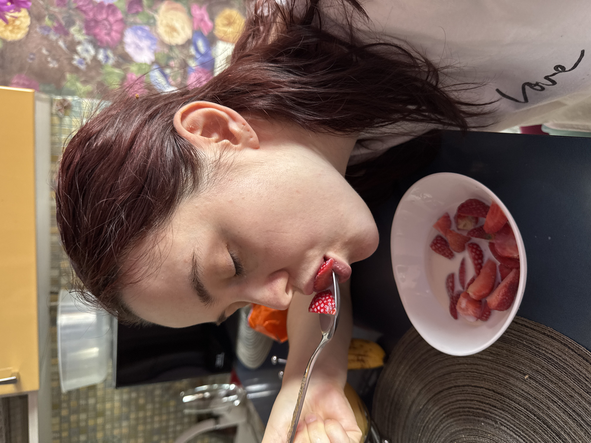 A person is shown in a close-up shot, eating strawberries and cream from a white bowl. The person has reddish-brown hair and is wearing a white shirt with the word "Love" written on it. They are holding a silver spoon with a strawberry in their mouth. The bowl of strawberries and cream is on a dark blue surface. In the background, there is a blurred view of a kitchen with a floral-patterned wallpaper. The photo was most likely taken indoors. There is nothing in the image to indicate it was taken in Sochi, Russia.