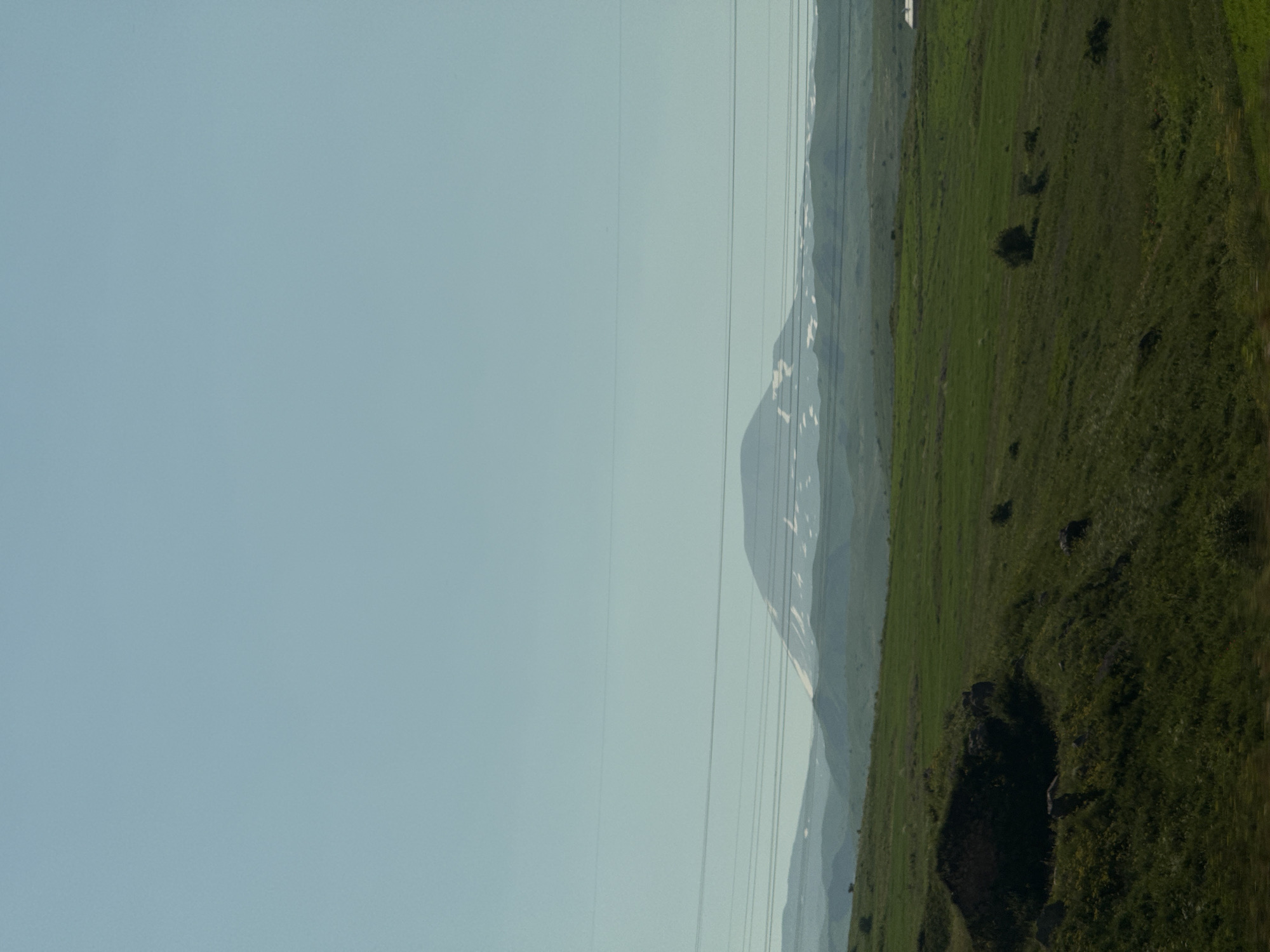 The image depicts a wide, open landscape under a clear, pale blue sky. In the foreground, a rolling green hill slopes downwards from left to right, covered in lush grass with scattered dark rocks and clumps of vegetation. A larger, dark, overgrown mound or structure is visible on the left side of the foreground. Further into the mid-ground, the landscape transitions to more distant, undulating green hills. A prominent mountain range, partially covered in snow, forms the backdrop. Several thin, horizontal power lines stretch across the mid-section of the image, obscuring parts of the mountain range. The overall impression is one of a natural, rural environment. The location is identified as being in Charentsavan, Armenia.