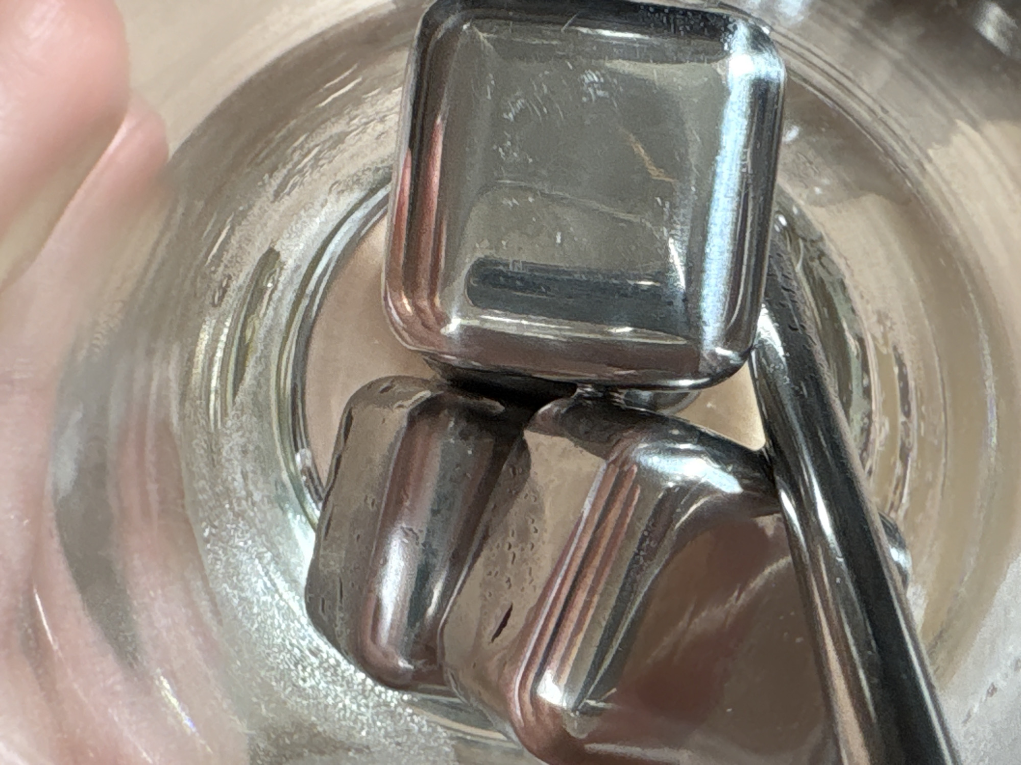 A close-up, overhead shot depicts a clear glass containing three metallic ice cubes and a metallic straw. The lighting is bright, causing strong reflections on the metallic surfaces. The scene is indoors, with a hand partially visible on the left edge of the frame. The glass appears to be empty, devoid of liquid. No specific location is identifiable.