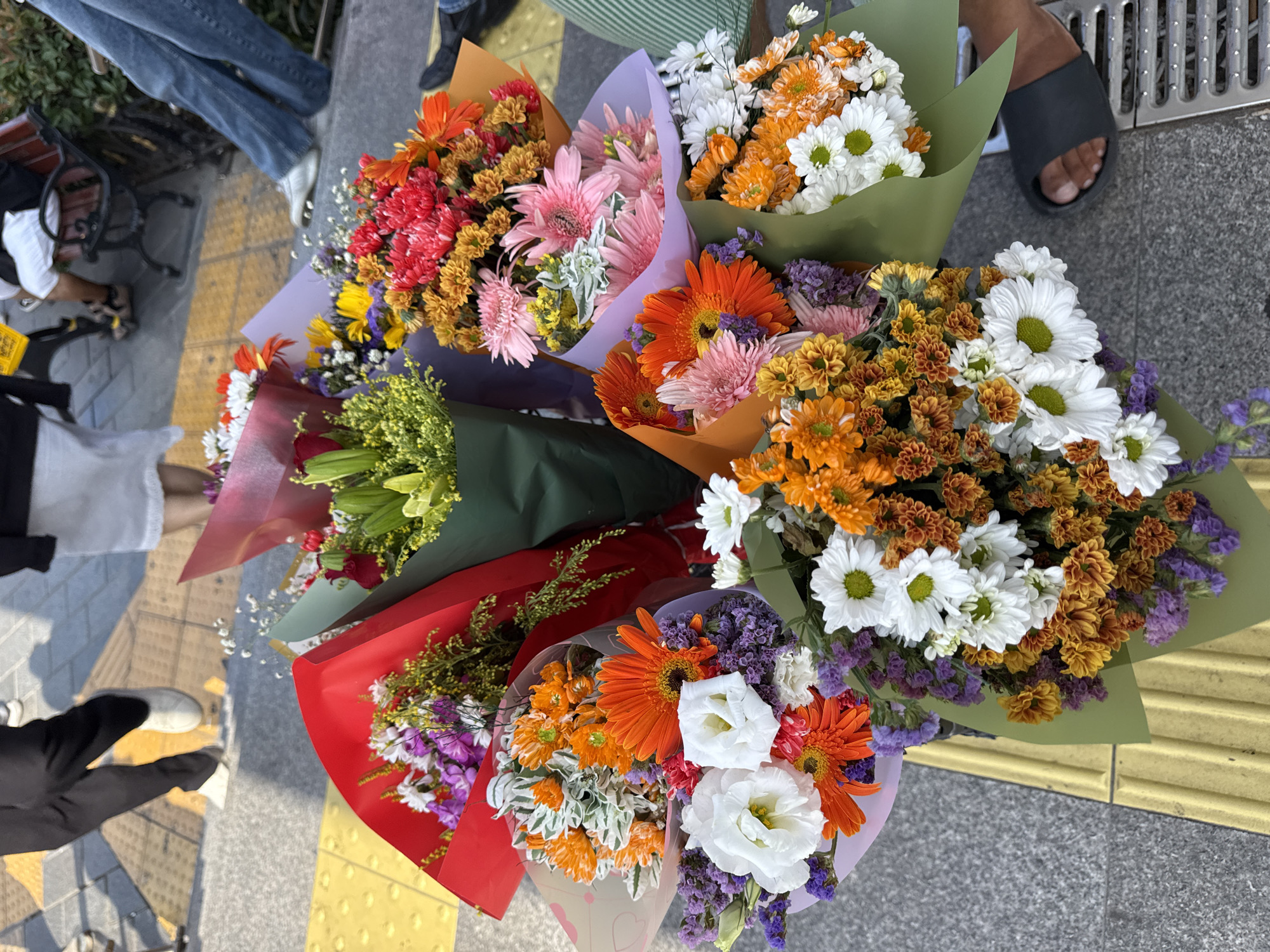 In Üsküdar, Turkey, a collection of vibrant floral bouquets is displayed on a gray, textured sidewalk with a prominent yellow tactile paving strip. The flowers, wrapped in various colored papers including red, green, purple, and orange, are arranged in a clustered fashion, suggesting they are for sale or have just been purchased. Among the blossoms are white daisies with green centers, bright orange gerbera daisies, pink gerbera daisies, red chrysanthemums, and purple statice. The background shows the lower legs and feet of several individuals. One person is wearing black trousers and white sneakers, another is in light-colored shorts and sandals, and another is in blue jeans and white sneakers. A dark-colored park bench is partially visible to the right. The scene conveys a bustling urban atmosphere with a focus on the colorful floral arrangements.
