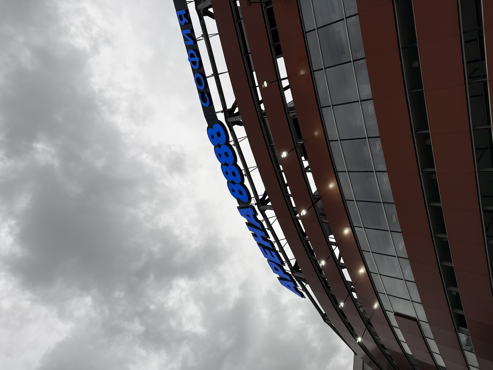 The image shows a low-angle view of a building with a sign that reads "ARENA SOFIA 88888". The sign is in blue Cyrillic letters, indicating the building's name and a possible phone number. The building has a modern architectural design with rows of windows and brown horizontal panels. The sky is overcast with heavy clouds. The location is Sofia, Bulgaria, as indicated by the Cyrillic text "София" on the sign.