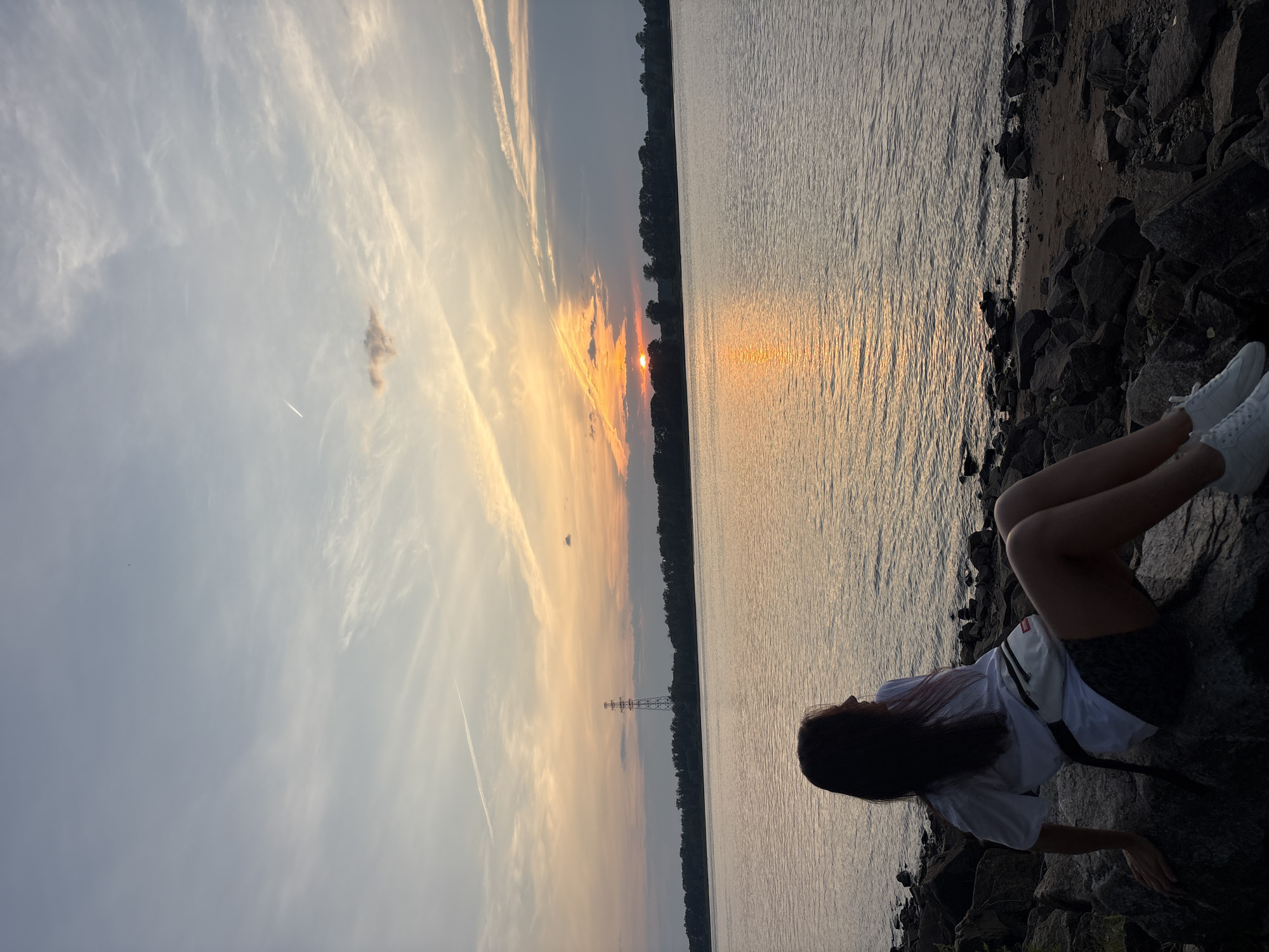 A young woman with dark hair, wearing a white t-shirt and patterned shorts, sits on a rocky shore facing a wide body of water. Her body is angled towards the left of the frame, with her head turned to the right, gazing at the sunset over the water. She has a white fanny pack around her waist. The water is calm, reflecting the warm colors of the sky. In the distance, a line of trees forms the horizon, and a tall, industrial tower stands prominently against the sky. The sky is filled with hues of orange, yellow, and blue, with scattered clouds and contrails from an airplane. The rocks in the foreground are dark and uneven, creating a natural border between the land and the water. The overall scene suggests a peaceful, contemplative moment during a sunset by a river.