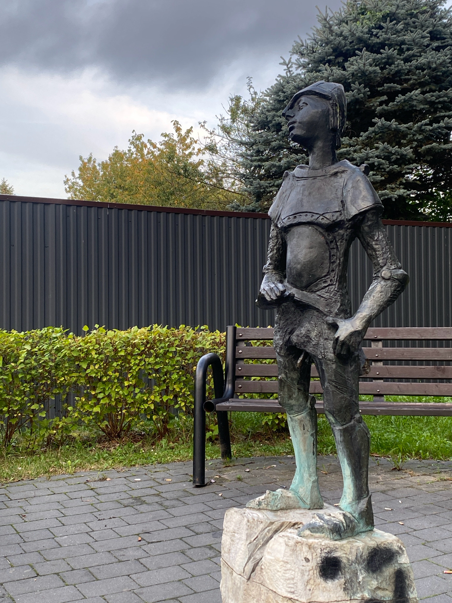 The image displays a bronze statue of a young person in a military uniform, standing on a rocky base. The statue is located in a park-like setting with a paved walkway, a wooden bench, and a hedge. A tall, dark fence forms the background, with trees behind it. The sky is overcast.
This statue, known as "The Young Soldier," is located at the intersection of Aleje Wolności and ul. Kazimierza Pułaskiego in Nowy Sącz, Poland. It was erected in honor of the young soldiers who fought in the defense of Nowy Sącz in 1939.