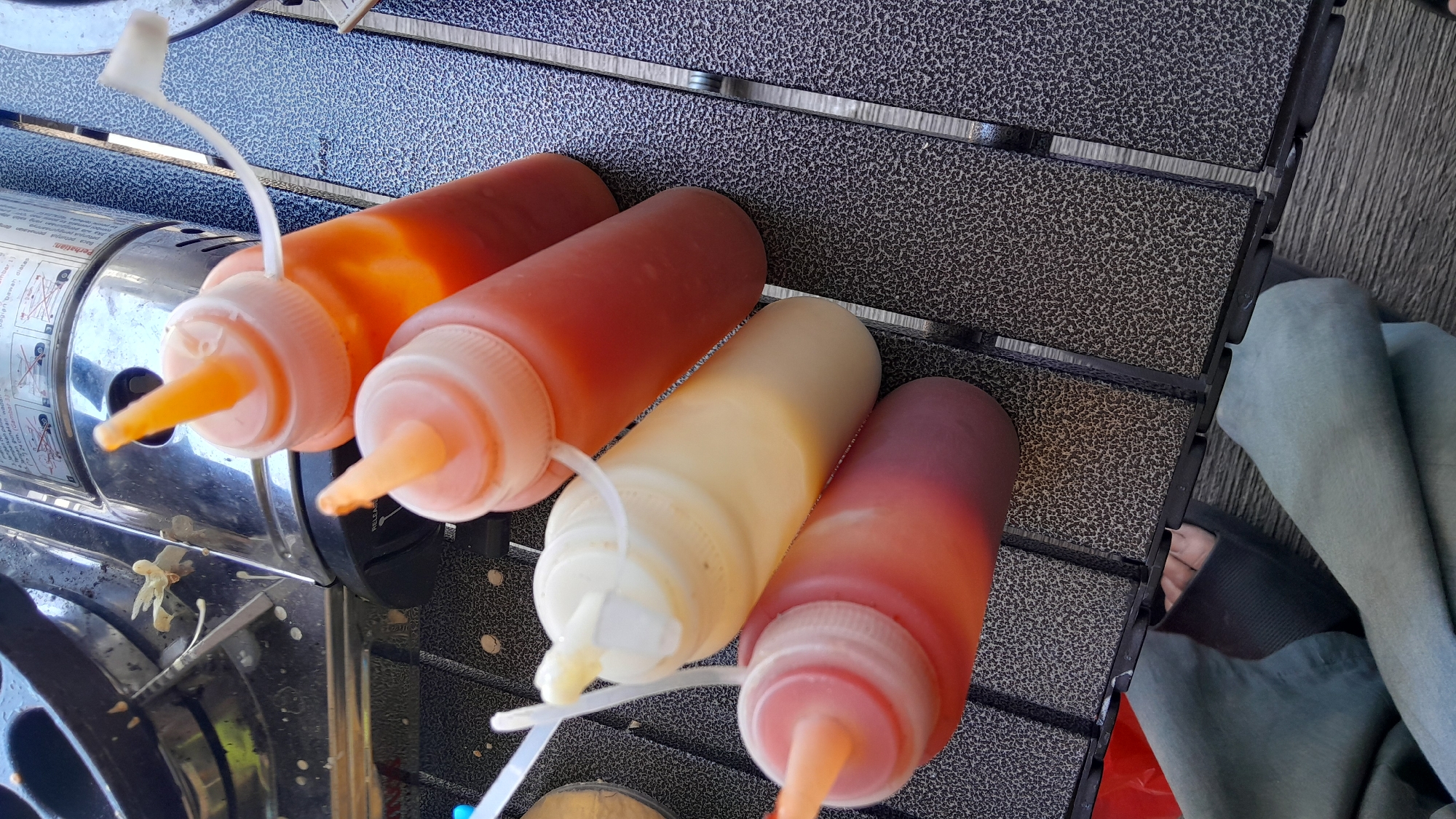 Four squeeze bottles containing sauces are arranged on a textured, dark grey table, which appears to be outdoors. The bottles are positioned diagonally, with their orange nozzles pointing upwards. From top to bottom, the visible sauces are: an orange-red sauce, a pinkish-red sauce, a creamy white sauce, and a darker red sauce at the bottom. A portable gas stove with some white residue on its surface is visible in the upper left quadrant of the image. In the lower portion of the image, a person's legs, clad in light blue pants, are visible, along with dark footwear. The overall setting suggests a casual outdoor meal or gathering. This scene could be in Bandung City, Indonesia, given the context provided, possibly at a picnic area or an outdoor food stall.