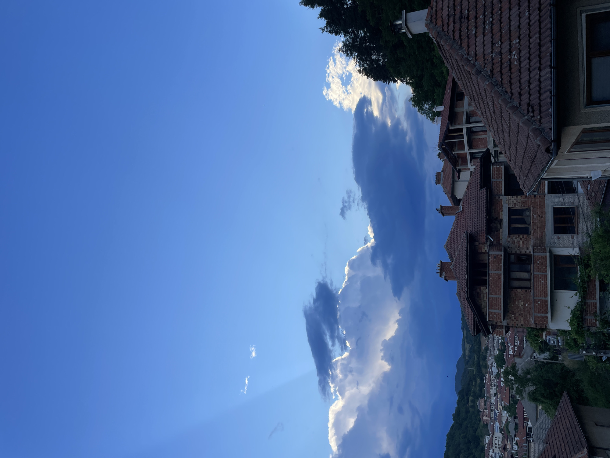 This is a vertically oriented, wide shot of a town in Zlatograd, Bulgaria. The scene is dominated by a vast, clear blue sky on the left, with a few wispy clouds scattered throughout. On the right, several buildings with terracotta tile roofs are visible, some appearing to be under construction with exposed brickwork. A dense line of trees runs along the top edge of the frame, partially obscuring the sky. The town below unfolds down a hillside, a collection of buildings with similar tiled roofs, nestled amongst lush green vegetation. The overall impression is of a sunny day, likely late afternoon or early evening, given the warm light on the clouds and the deep blue of the sky. There are no people or specific activities depicted, but the setting suggests a tranquil, possibly residential area.