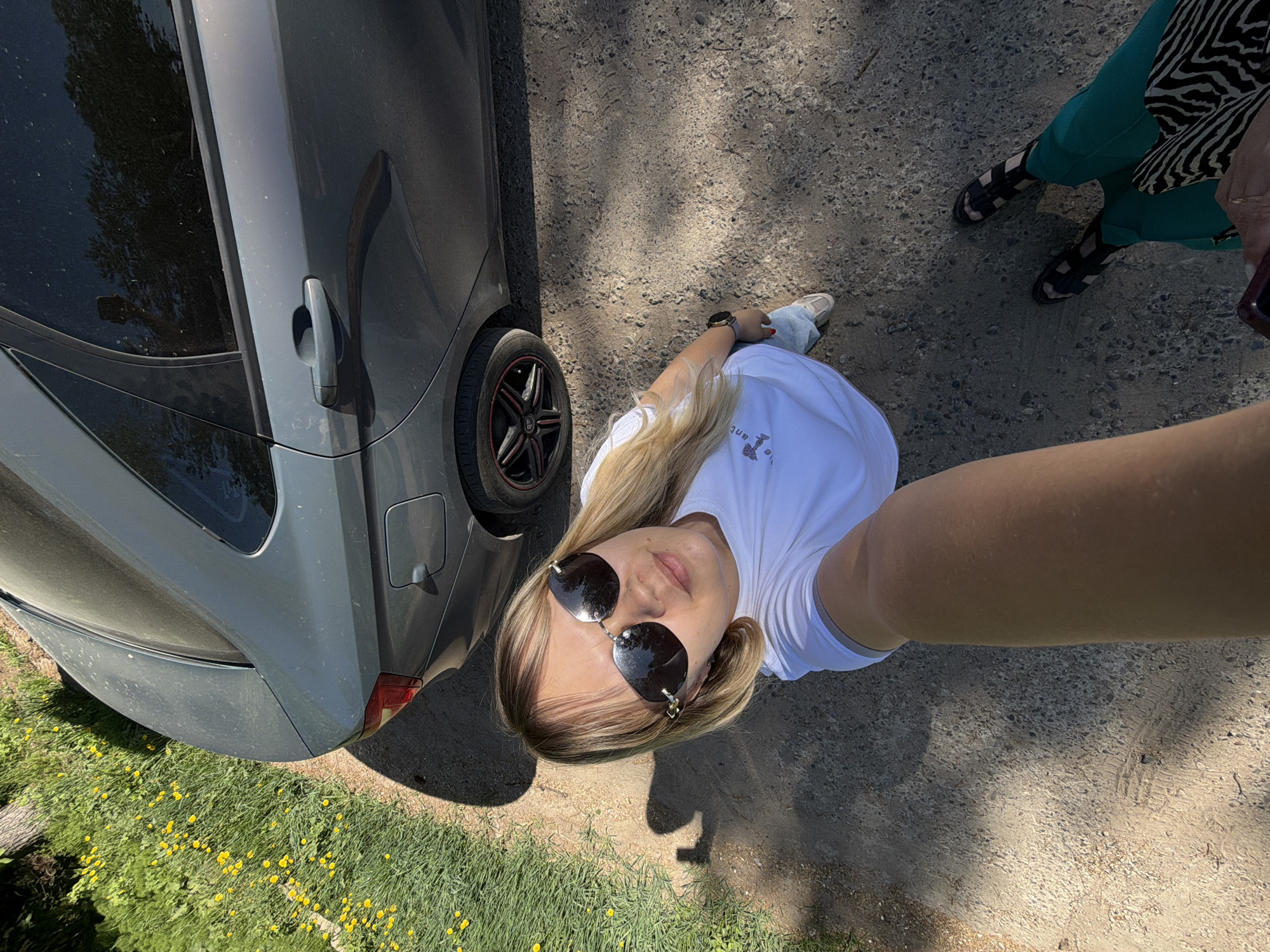 A woman is taking a selfie outdoors next to a gray car. She is wearing sunglasses and a white shirt. Another person wearing green pants and zebra print is partially visible in the upper right corner of the image. The location appears to be a dirt path or road next to a grassy area with yellow flowers. The selfie appears to be taken during daylight. It is taken in Chaikovsky, Russia.