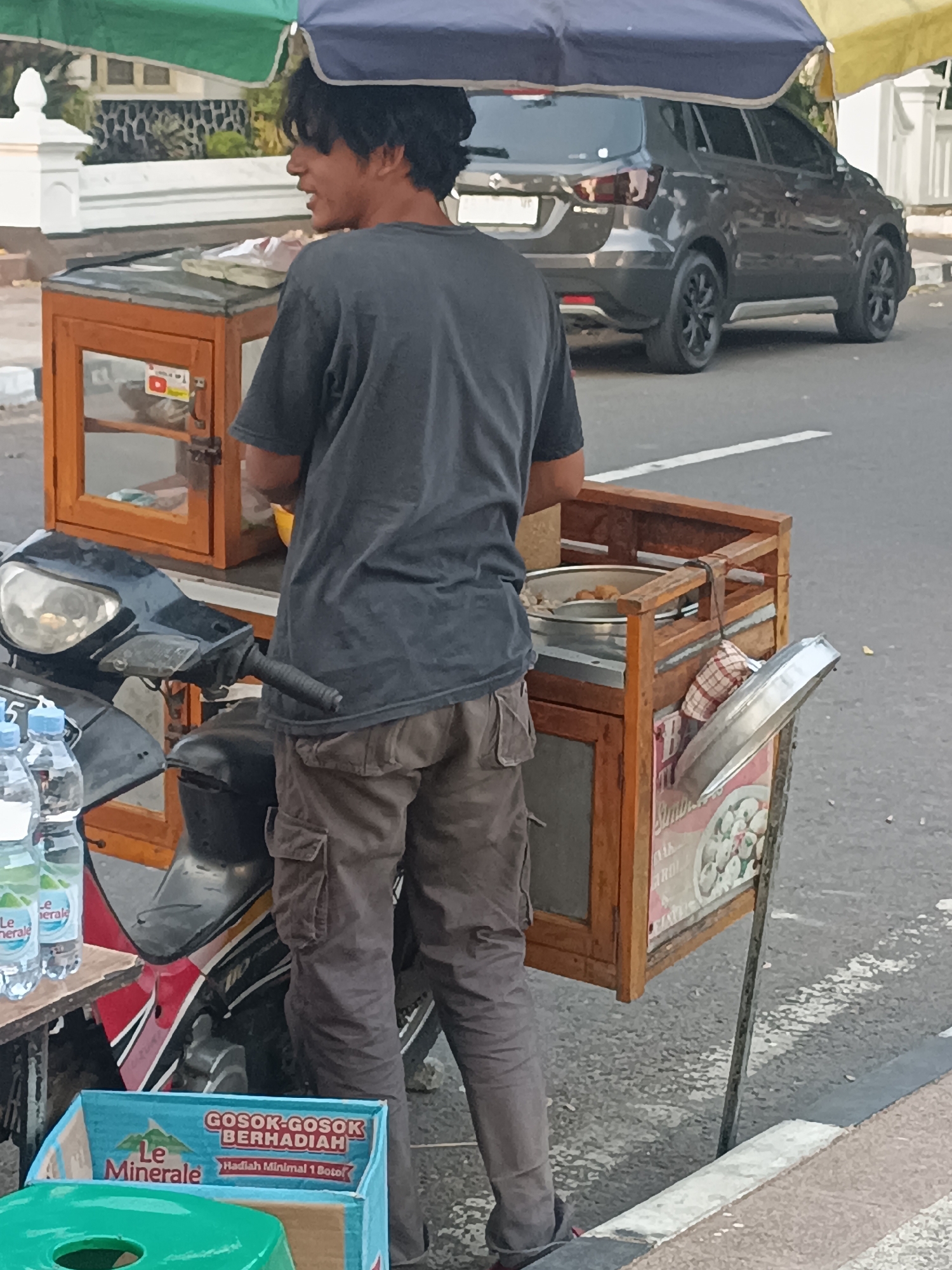 A young man with dark, messy hair stands behind a wooden food cart attached to a motorcycle in Sleman, Indonesia. He is wearing a dark grey t-shirt and loose-fitting, tan cargo pants. The food cart has a glass display case on the left and a larger open section with a metal bowl containing food on the right. A checkered cloth is hanging from the right side of the cart. Next to the motorcycle, on a small table, are two bottles of "Le Minerale" water. In the background, a dark grey SUV is parked on the side of the road. The scene is captured from a slightly elevated perspective, looking down at the man and his mobile food stall. The environment suggests an outdoor urban setting, possibly a roadside market or street food area.