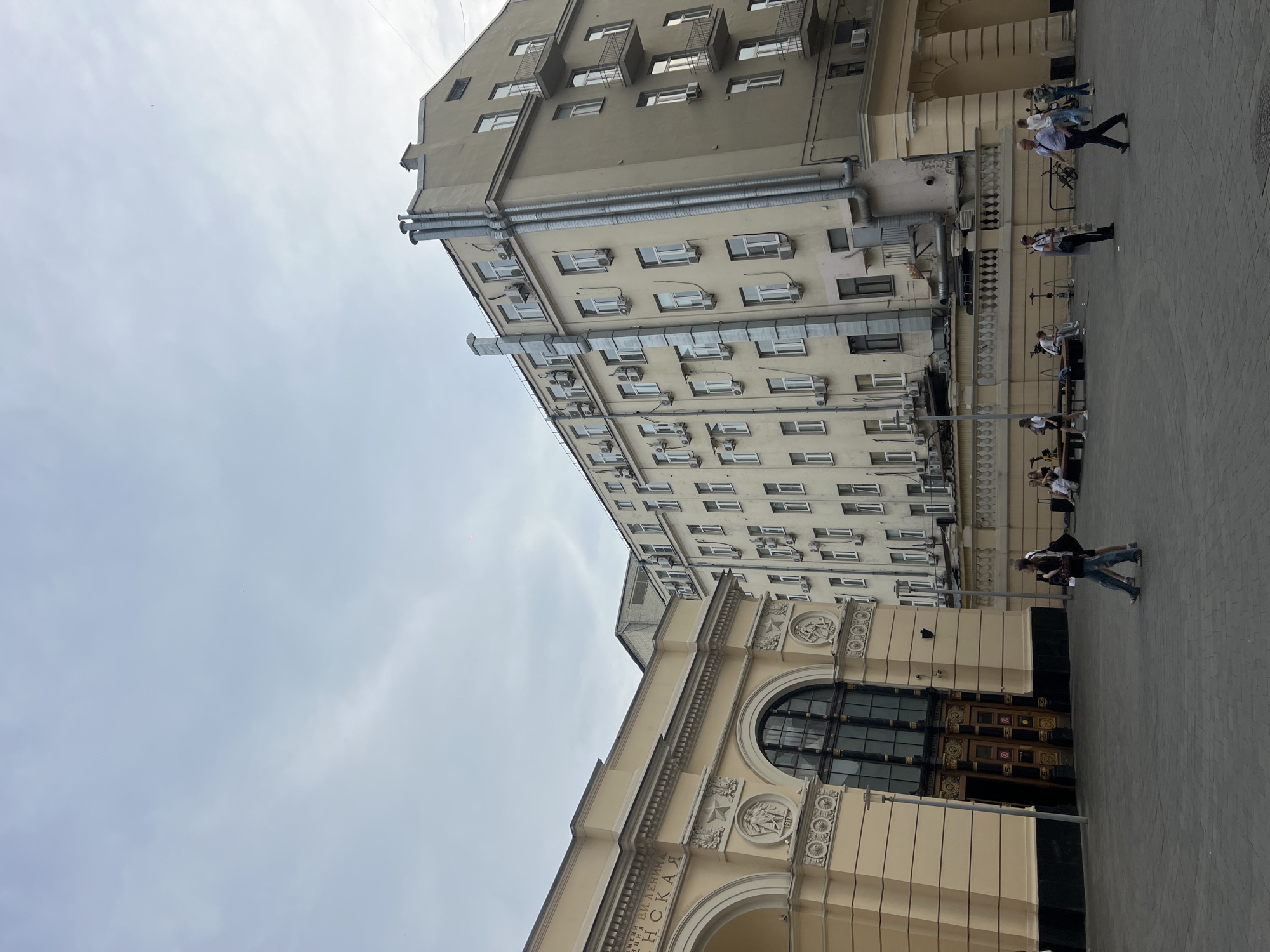 The image depicts a street scene in Moscow, Russia, in front of a building that appears to be a cultural institution or historical landmark. The architecture of the main building on the left is ornate, with arched entrances and decorative carvings, and bears Cyrillic inscriptions that suggest it is a significant establishment. To the right, a taller, more modern apartment building rises, characterized by numerous windows and external air conditioning units.
Several people are visible in the foreground and mid-ground, engaged in various activities. Some are walking, while others are seated on benches or standing around. The ground is paved with grey cobblestones or tiles. The sky is overcast with clouds, indicating a temperate day. The overall impression is one of an urban environment with a mix of historical and contemporary architectural styles.