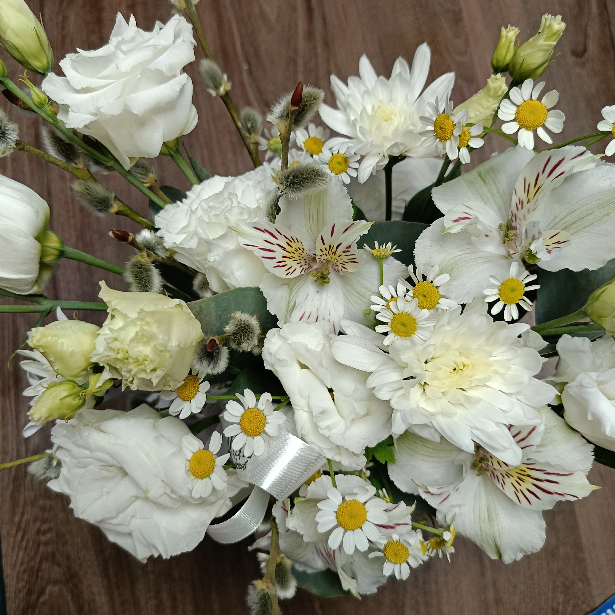 A close-up, eye-level shot depicts a bouquet of white flowers with yellow centers arranged on a brown wooden surface. The flowers include roses, chrysanthemums, daisies, and alstroemeria. Sprigs with fuzzy, silver-gray buds are interspersed among the blooms. A silver ribbon is visible near the lower left corner of the bouquet. The location is Markovskii, Russia.