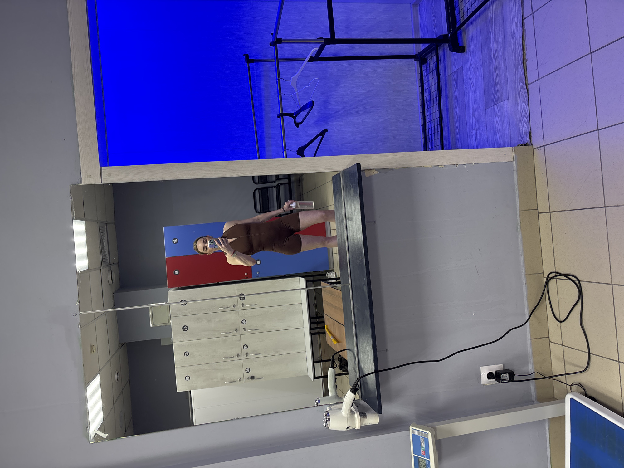 The image depicts a changing room or locker room, likely within a gym or fitness center. A person, appearing female, is the central subject, taking a selfie in a large, rectangular mirror. She is wearing a form-fitting, dark brown sleeveless jumpsuit and holds a clear water bottle in one hand and a smartphone in the other, which is used for the selfie. Her reflection shows her light-colored hair pulled back.
Behind the person in the reflection are two sections of lockers. One section features light gray, wood-grain style lockers with silver handles, displaying numbers such as 10, 11, 12, 13, 14, 15, and 16. Another section contains brightly colored blue and red lockers, with numbers 32 and 33 visible. The ceiling reflected in the mirror has multiple fluorescent strip lights.
In the foreground, beneath the mirror, a dark counter holds two white hair dryers plugged into a wall outlet, with black cables neatly arranged. A white digital scale with a blue display, partially showing "MEDISANA," is also visible. The floor is covered with light-colored tiles.
A distinct area in the upper right part of the scene (behind the mirror's perspective) features a dark metal clothes rack with several white and black hangers. This section is dramatically illuminated by vivid blue ambient lighting, contrasting with the brighter, more functional lighting of the changing area. The walls are a light gray. The water bottle held by the person has "Prime One" printed on it.