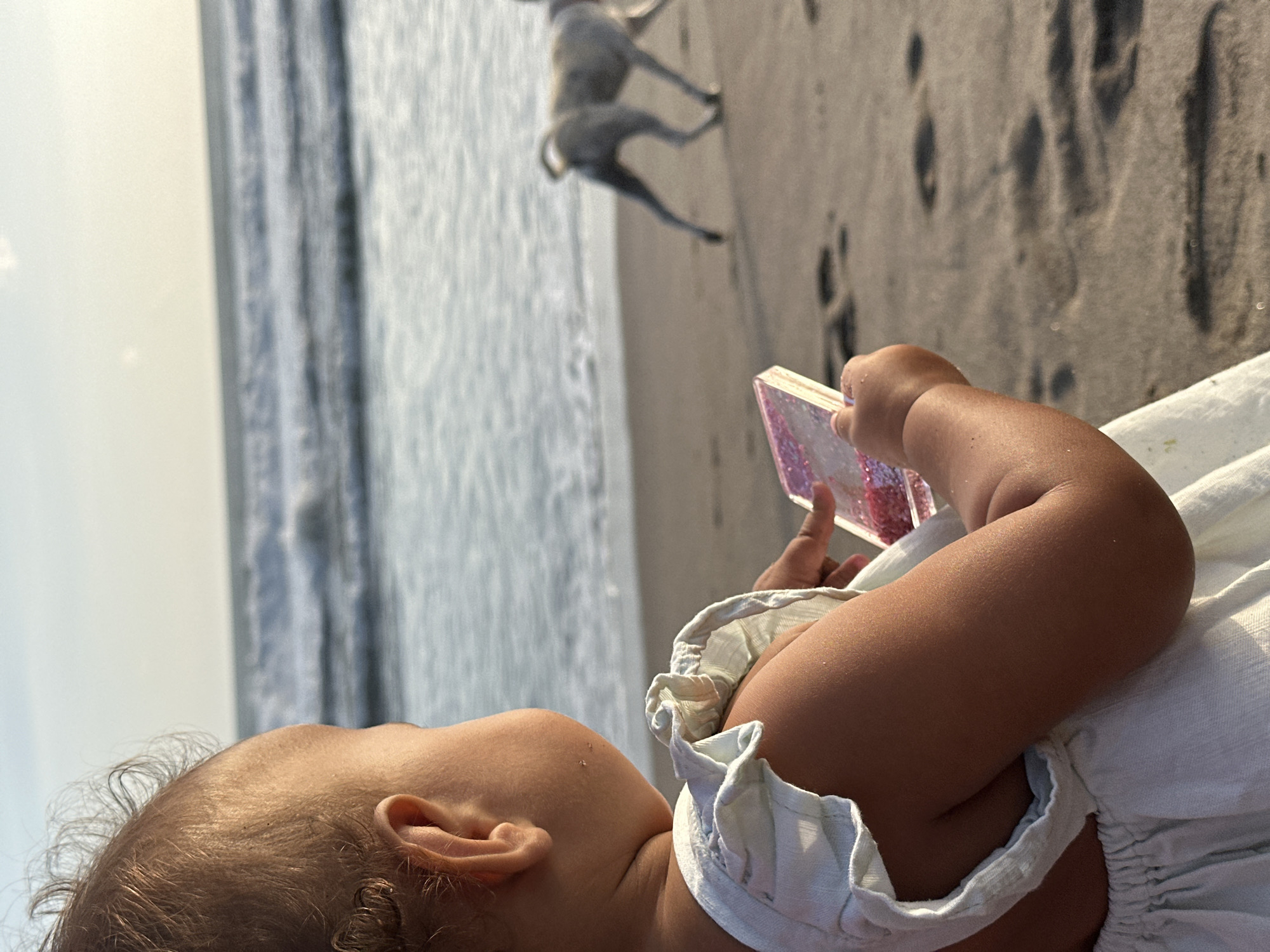An infant wearing a light blue dress sits on a sandy beach in North Kuta, Indonesia, facing away from the camera and towards the ocean. The child is holding a small, rectangular object with pink glitter. The ocean has moderate waves, and the sky is bright and clear, suggesting it is either early morning or late afternoon. In the background, a light-colored dog runs along the shoreline. Footprints are visible in the sand. The overall lighting indicates a warm, sunny day.