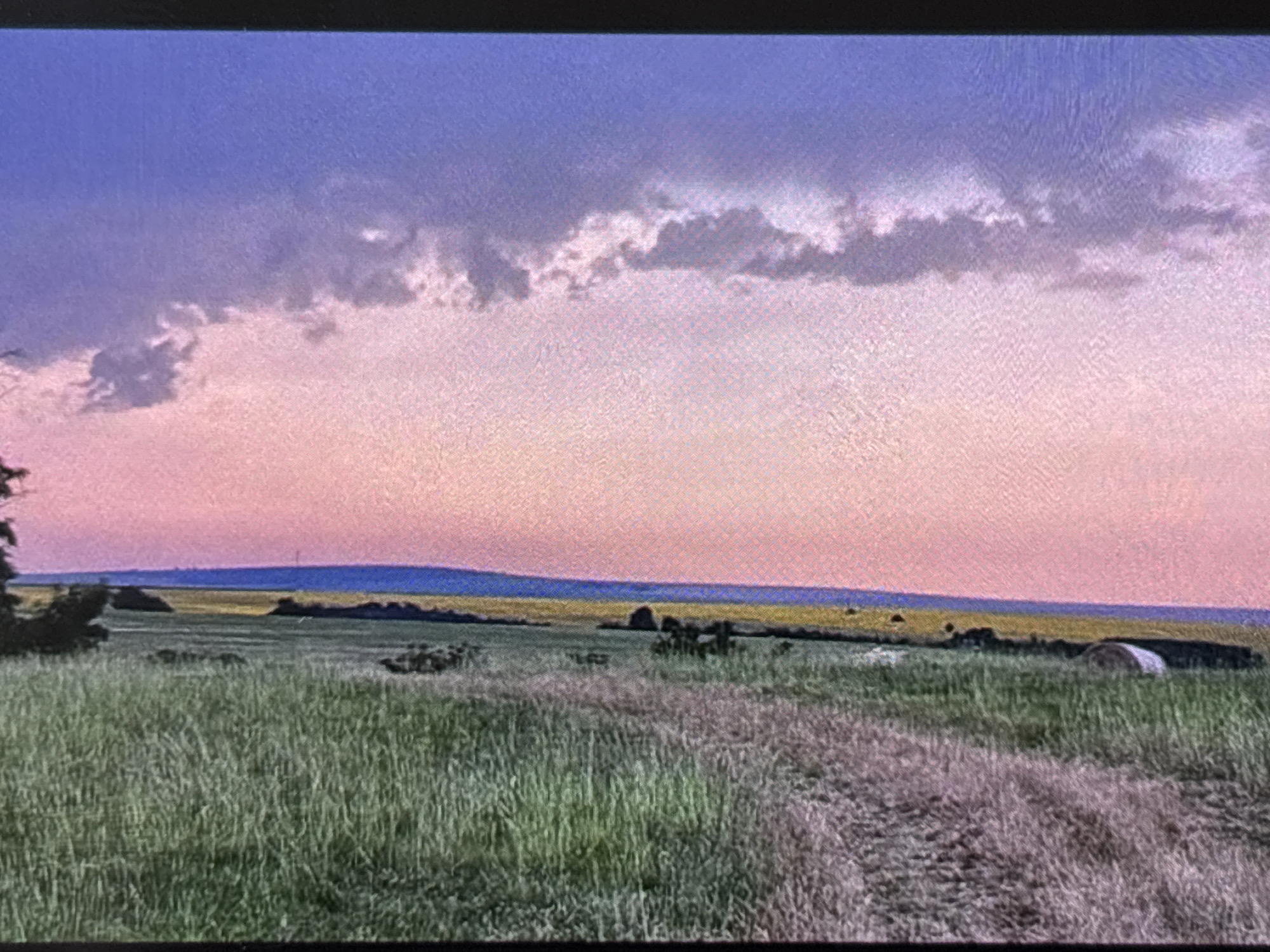 This media file captures a serene rural landscape, likely at sunrise or sunset, located near Wolverhampton, United Kingdom. The scene features expansive fields under a colorful twilight sky.
The sky transitions from a muted blue-purple at the top, down to a soft pink and orange gradient near the horizon. A prominent band of textured, darker clouds stretches horizontally across the mid-section of the sky.
Below the sky, the distant landscape reveals a thin strip of deep blue or purple land, possibly distant hills or another type of field. Beneath this, a wider field of vibrant yellow is visible, suggesting a crop like rapeseed in bloom.
The midground and foreground are dominated by a lush green field, likely grass or a young crop, with varying textures and heights. A dirt track or path with visible ruts winds through this green field, entering from the lower right and extending towards the center. To the far right in the midground, a large round hay bale rests in the field. On the left side, a cluster of dark trees or bushes partially frames the view.
The overall mood is peaceful and tranquil. There are no people visible, nor is any text present in the image. The photograph appears to be an image of a screen, as evidenced by a visible pixelation or screen-door effect throughout.
