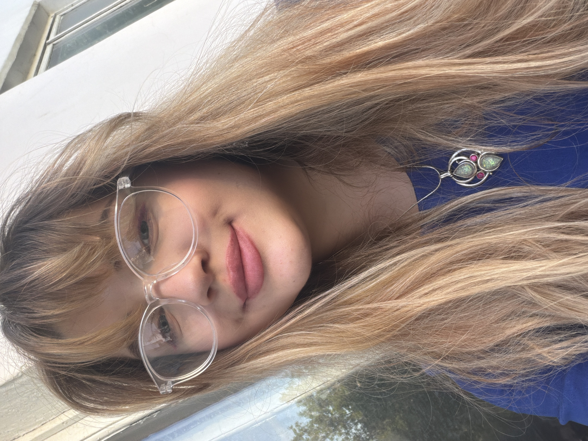 A woman with long, blonde hair and clear-framed glasses is shown in a selfie-style photograph. She is wearing a blue shirt and a pendant necklace with green and pink gemstones. The background includes a window with a white frame and what appears to be an exterior view with greenery. The exact location in Naberezhnye Chelny, Russia, is not discernible from the visual information provided.