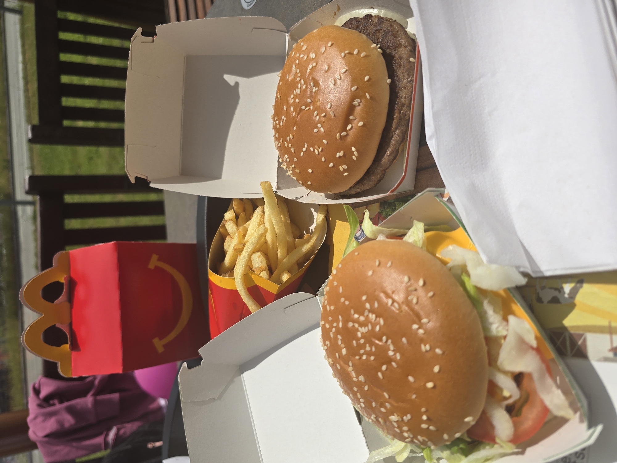 An overhead outdoor shot captures a McDonald's fast food meal set on a dark table, likely in Erding, Germany, under bright daylight. The scene features a red McDonald's Happy Meal box with a golden arch logo and a yellow smiley face on its front, positioned towards the upper left. Adjacent to it, a red and yellow carton of French fries is partially visible, containing numerous golden-brown fries. To the right, two open white cardboard burger boxes are present. The box on the far right holds a plain hamburger with a sesame seed bun and a beef patty. The box on the left contains another hamburger, also with a sesame seed bun, topped with lettuce, a tomato slice, and a slice of cheese over a beef patty. White paper napkins or wraps are visible towards the bottom right. In the blurred background, dark brown wooden fence posts or chair backs are discernible, with green grass beyond them and a faint outline of a road or path. A blurred pink object and a folded purple garment are partially visible in the distant left background. No individuals are depicted, but the arrangement suggests a meal ready for consumption.