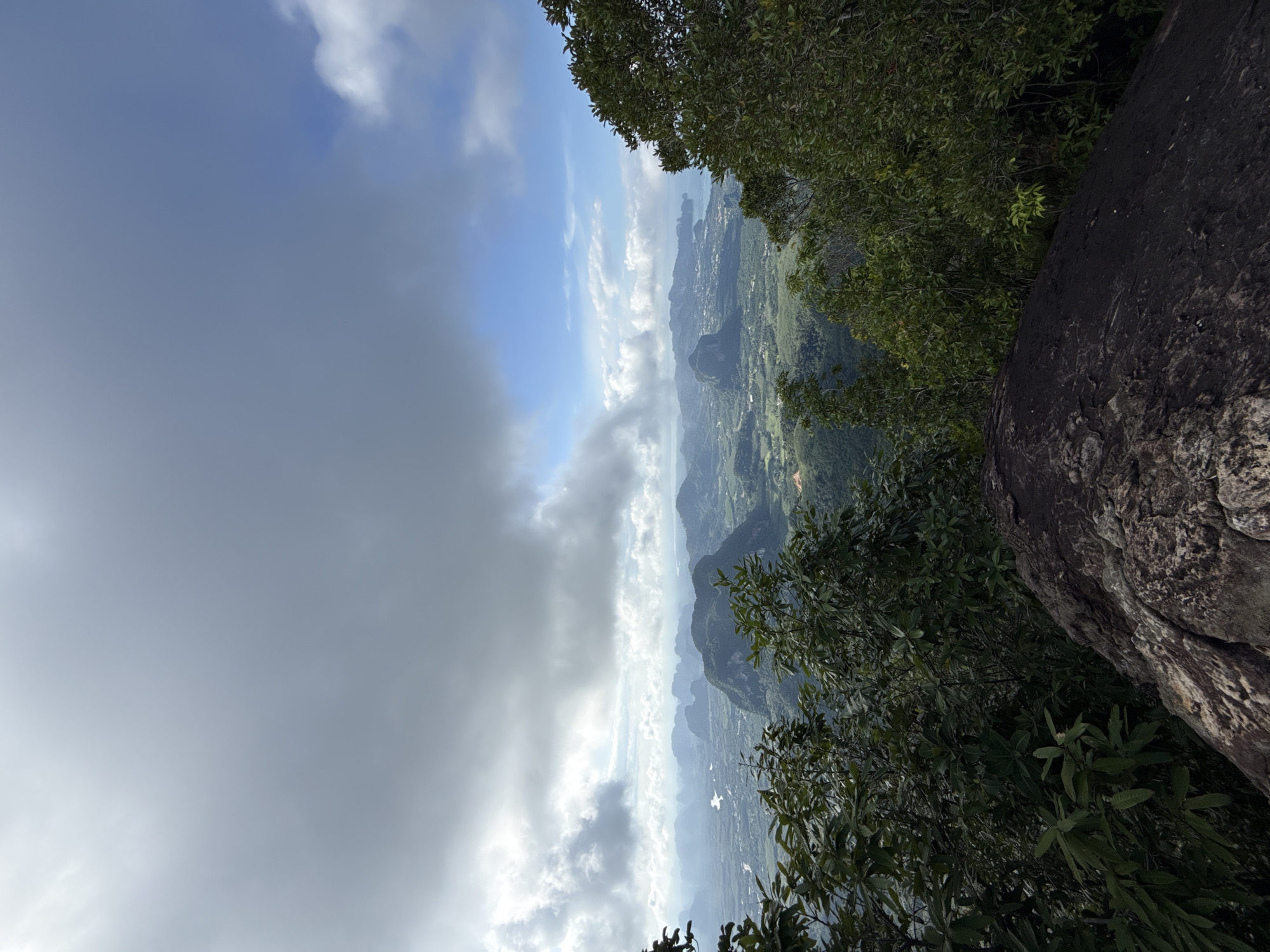 This is a landscape photograph taken from a high vantage point, overlooking a valley with karst formations and a coastline. The foreground is dominated by dark, textured rocks and lush green foliage from trees. In the middle ground, a sprawling valley unfolds, dotted with smaller hills, vegetation, and what appears to be a river or waterway. Further in the distance, more prominent, rugged karst mountains rise, some with visible structures or settlements at their bases. The horizon reveals a hazy coastline and the ocean. The sky above is a mix of dramatic, dark clouds and patches of clear blue sky with fluffy white clouds. The scene suggests a natural, tropical environment, likely captured during or after a period of rain due to the overcast conditions and potential haze. The location is identified as Ban Din Daeng Nui, Thailand, suggesting this viewpoint is from a mountain or elevated area within or overlooking this region. No people or animals are visible in the image.