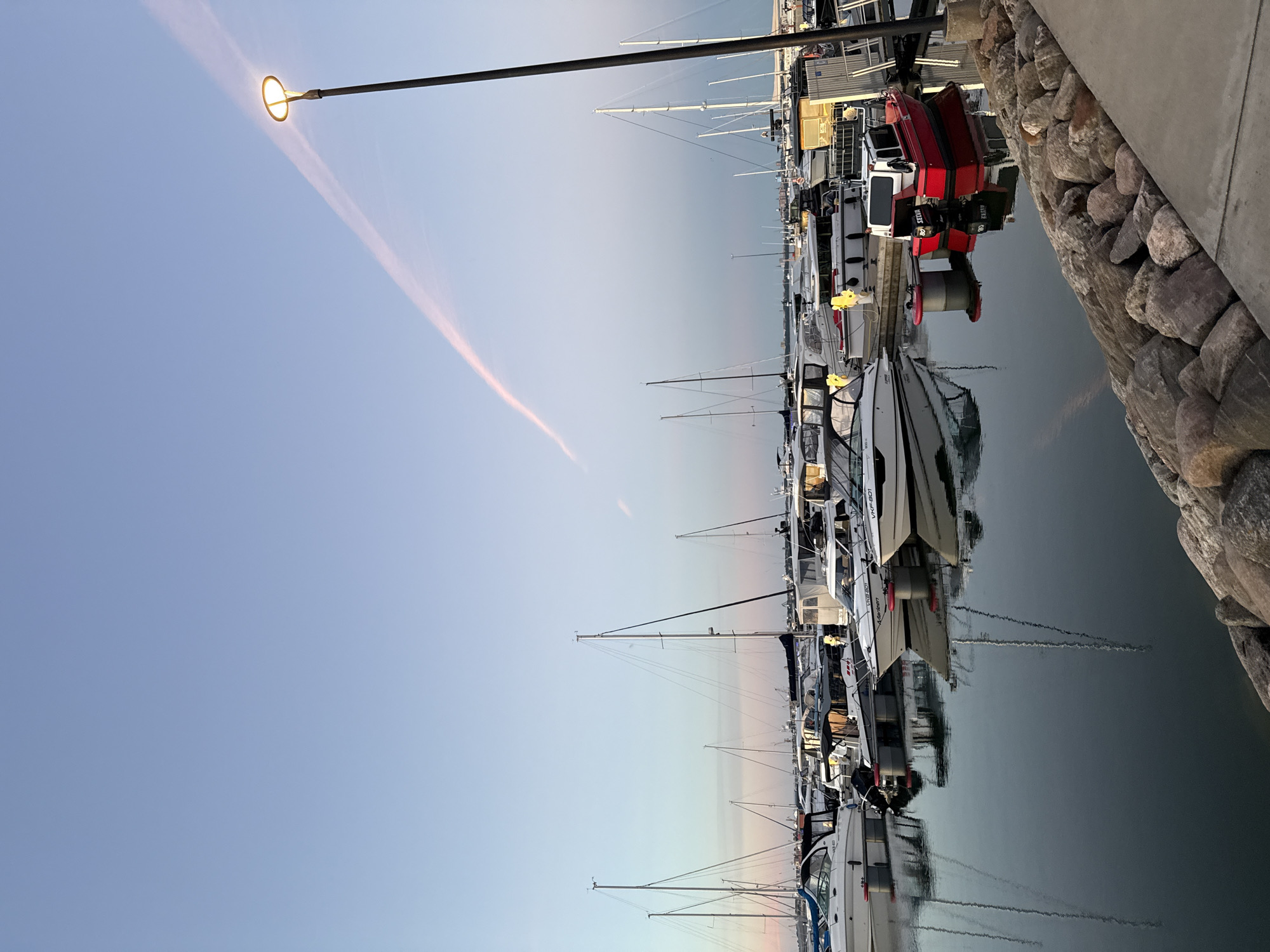 This is a photograph of a marina in Tallinn, Estonia, taken during twilight. The sky is clear and a pale blue with a faint pink contrail stretching across it. A street lamp is illuminated on the right side of the frame. The marina is filled with various boats, including sailboats and motorboats, docked in neat rows. The boats are reflected in the calm, dark water of the harbor. In the foreground, a concrete walkway is lined with large, rough-cut rocks. The scene is peaceful and serene, with the twilight hour casting a soft glow over the water and boats.