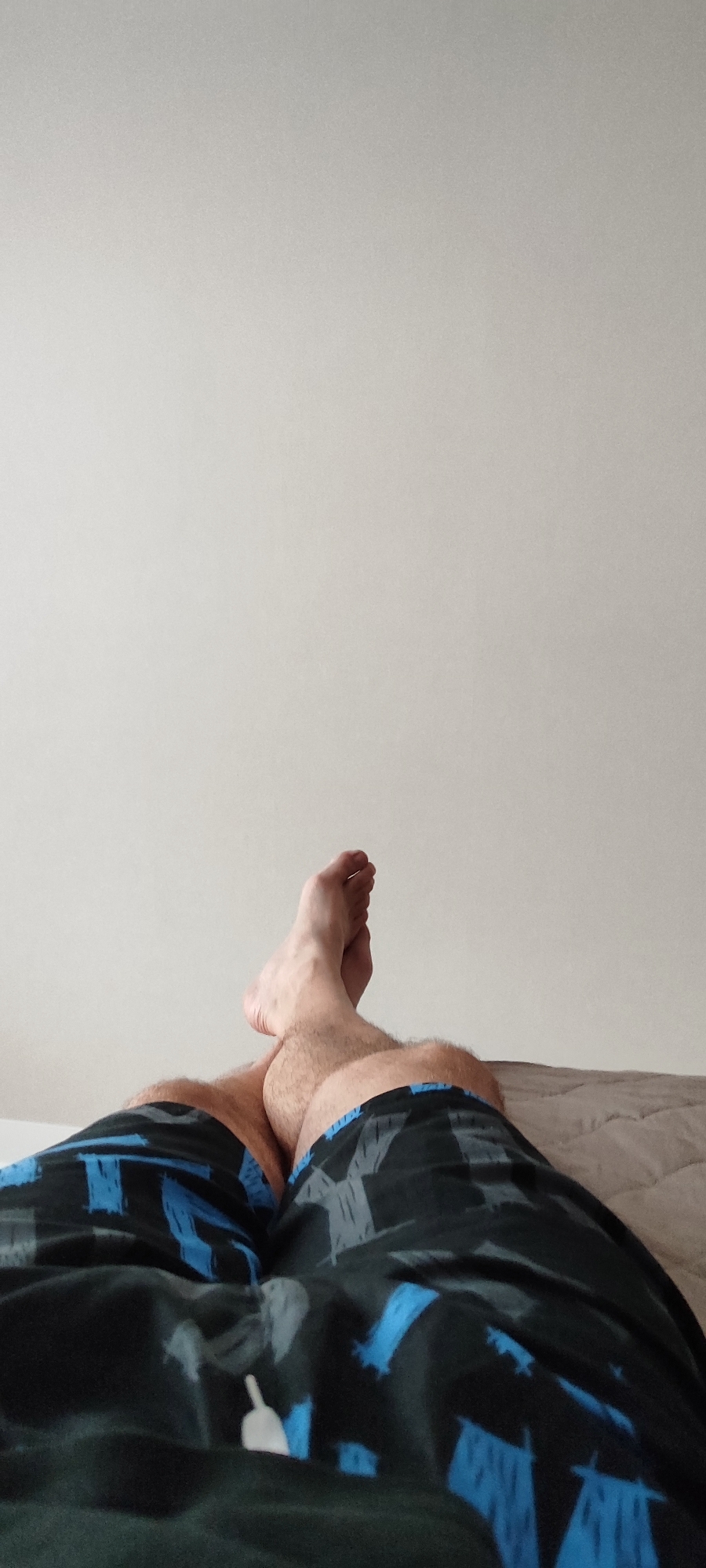 The image shows a low-angle, close-up view of a person's legs and feet, resting on a bed. The person is wearing black shorts with a blue and gray abstract pattern. The legs are crossed, with the left leg resting on top of the right. The feet are bare and visible, with the toes of the left foot pointing upwards. The background consists of a plain, off-white wall and a textured, light brown bedspread. The lighting suggests an indoor environment, likely a bedroom, with a soft, diffused light source. The focus is sharp on the legs and feet, with the background slightly blurred.