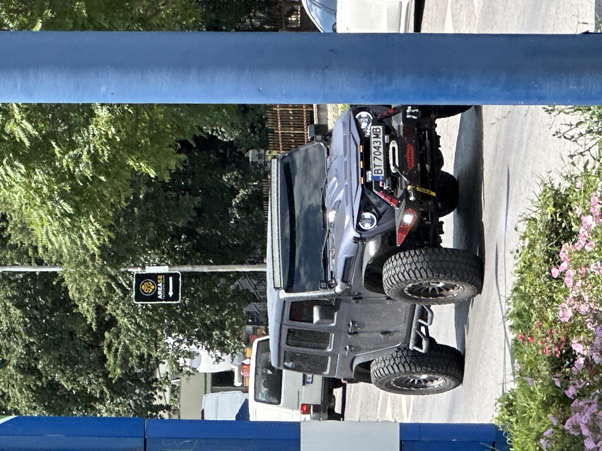 This image shows a close-up, vertically oriented view of a gray Jeep Wrangler parked outdoors. The Jeep is the primary subject, with its front grille, headlights, winch, and large, knobby tires clearly visible. The license plate reads "BT 7043 MB". A black and yellow sign with an emblem and an arrow pointing left, reading "AREA52", is also visible, mounted on a metal pole to the left of the Jeep.
In the background, a white van is partially visible behind the Jeep. Lush green trees with sunlight filtering through them dominate the upper left portion of the image. To the right, there is a bush with pink flowers and green foliage. A grey paved surface, likely a road or parking lot, forms the ground. The scene appears to be set during the daytime, with bright sunlight casting shadows. The overall mood is casual and outdoor-oriented. The presence of the sign "AREA52" might suggest a specific location or themed area. The vehicles and signage imply an urban or semi-urban setting in Bulgaria, possibly Veliko Tarnovo as indicated by the license plate prefix.