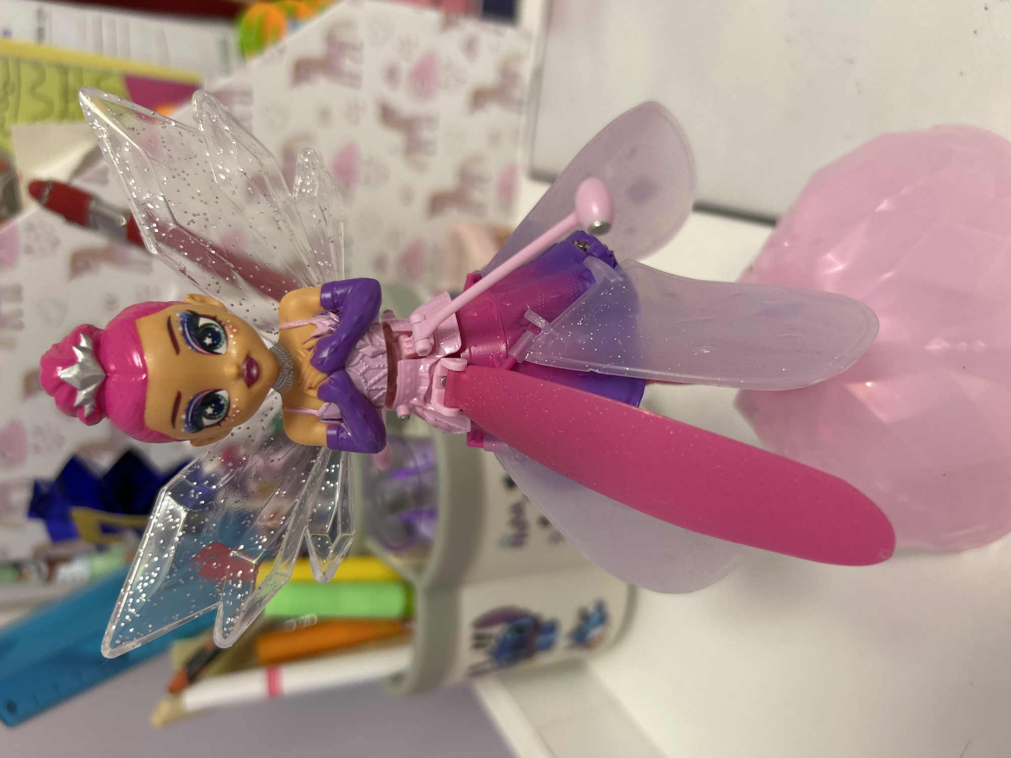 A close-up, eye-level shot captures a pink-haired fairy doll with purple gloves and a silver tiara. The doll has blue eyes and is posed with its hands clasped at its chest. It features translucent, glitter-speckled wings and a layered skirt in shades of pink and purple. The doll stands on a pink, faceted base. In the background, out of focus, are various desk supplies including pens and markers in cups, a blue ruler, and a white surface with a pink, unicorn-patterned wallpaper. The location of Burgas, Bulgaria is not present in the image.