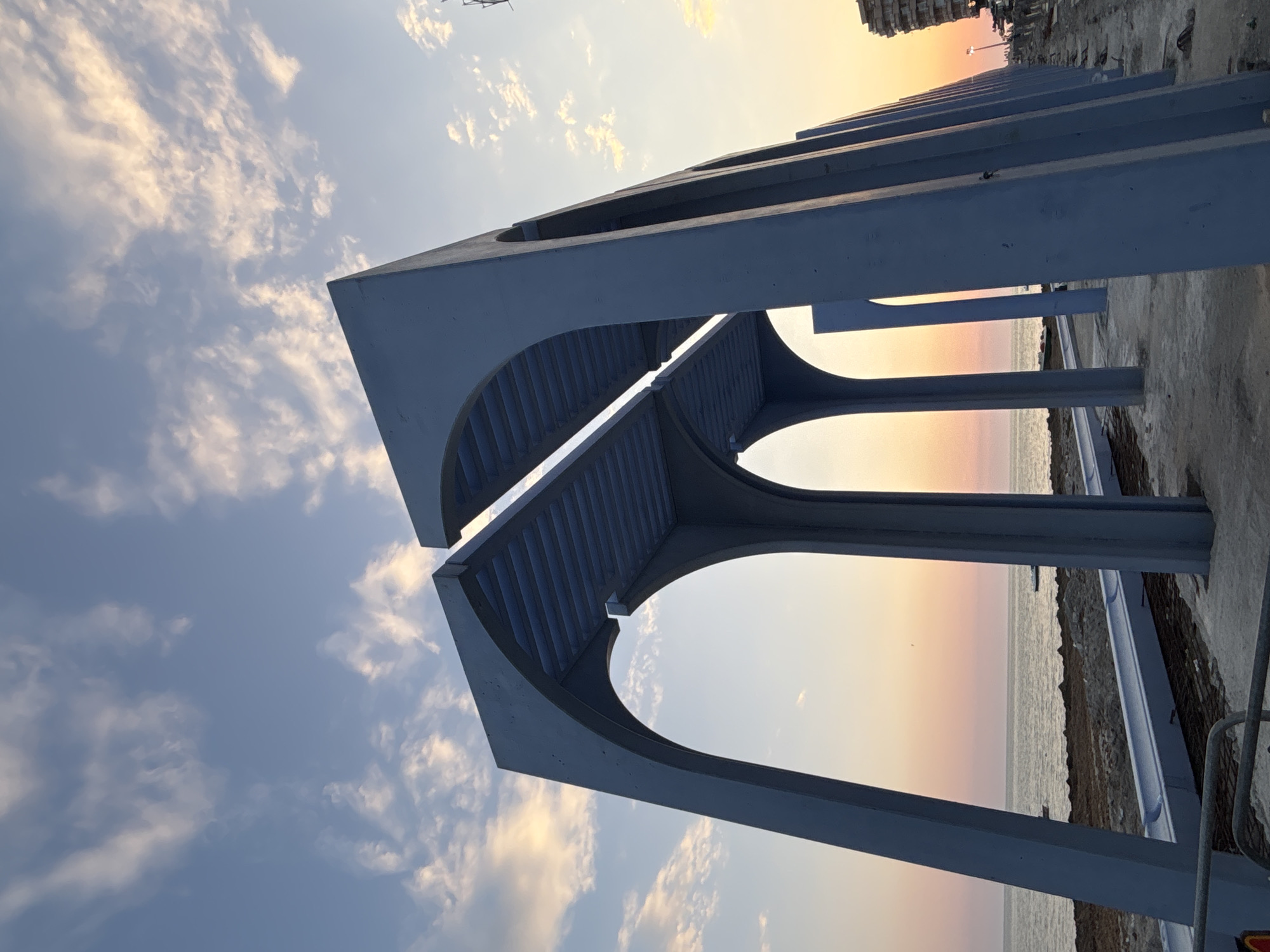 The image depicts a seaside scene in Bashkia Durrës, Albania, during sunset. A blue, modern architectural structure with arched openings and a slatted roof dominates the foreground. The structure appears to be a walkway or pavilion, with its construction possibly ongoing as indicated by visible materials on the ground.
The sea stretches out to the horizon, reflecting the soft hues of the setting sun, which casts a gradient of pink and orange across the sky. Scattered clouds with golden edges punctuate the blue sky. The shoreline consists of sand and some debris.
In the distance, along the coastline, a multi-story building is visible, suggesting a more developed area further down the promenade. There are no people or animals clearly discernible in the image. The overall impression is one of an outdoor public space at the end of the day, with a focus on the striking architectural element against the natural backdrop of the sea and sky.