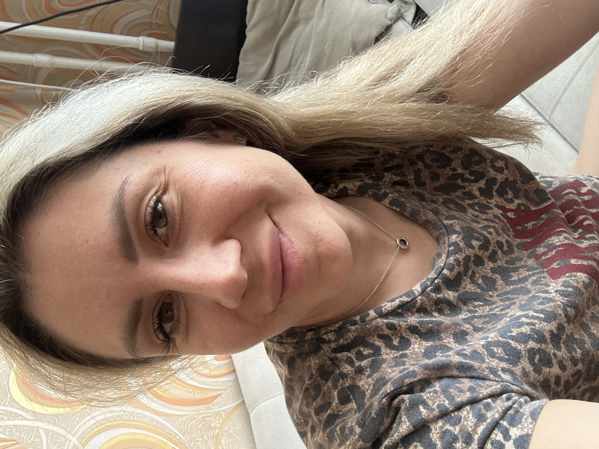 A close-up, waist-up selfie shows a woman with blonde hair and brown eyes. She is smiling and looking at the camera. She is wearing a leopard-print t-shirt and a silver necklace with a circular pendant. The background is out of focus, but appears to be a room with patterned wallpaper and furniture. The image was taken in Novoaltaysk, Russia.