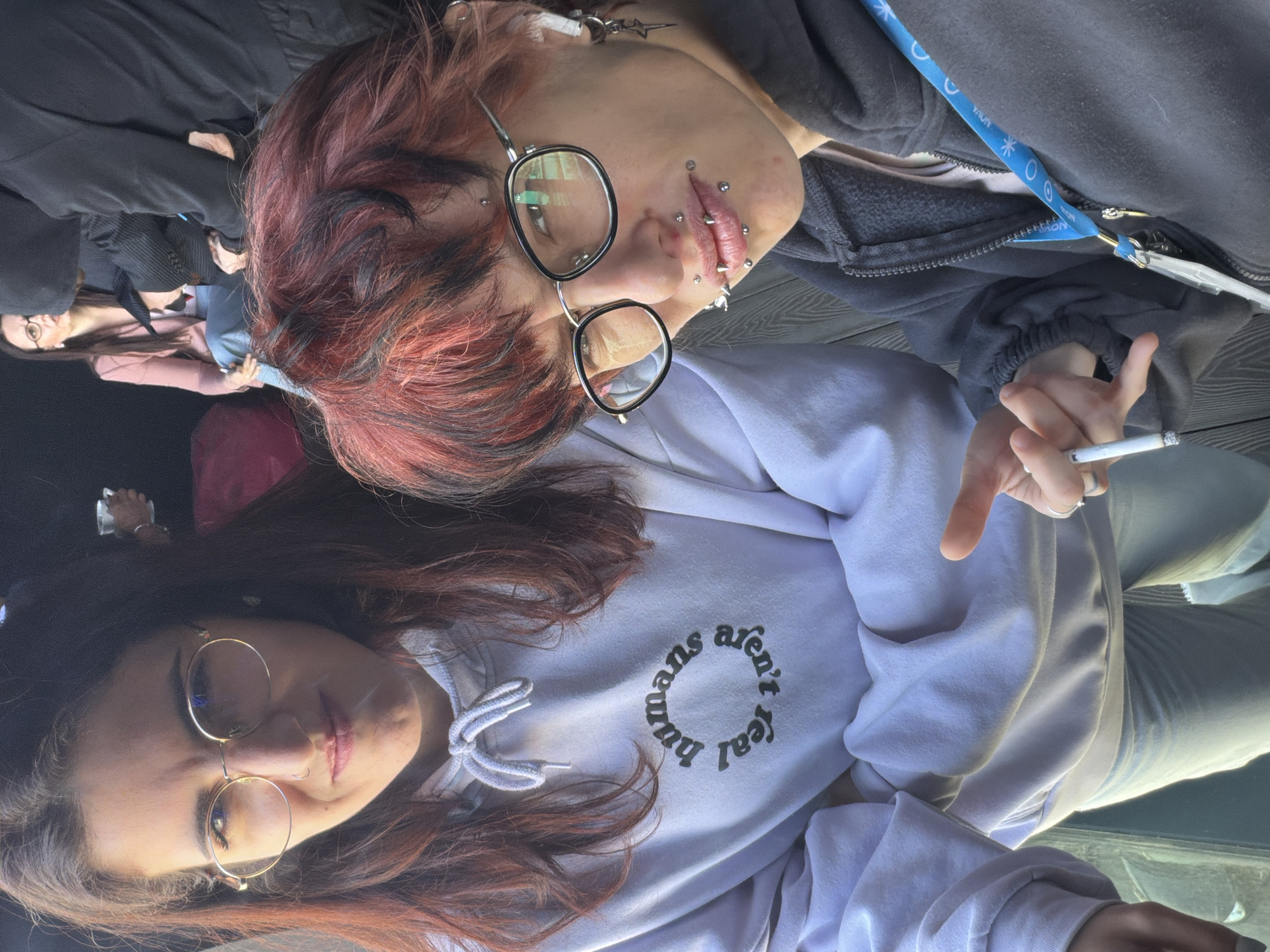 Two people are photographed from a high angle. The person on the left has long brown hair with a hint of red, round glasses, and is wearing a white hoodie with the words "humans aren't real" printed on it. The person on the right has short red and black hair, round glasses, and multiple piercings around their face. They are holding a lit cigarette. The location appears to be a high vantage point within a building, possibly a rooftop or high floor. Other people, including one wearing a pink top, are faintly visible in the background on the left side.
I am unable to identify the specific location in Sofia, Bulgaria.