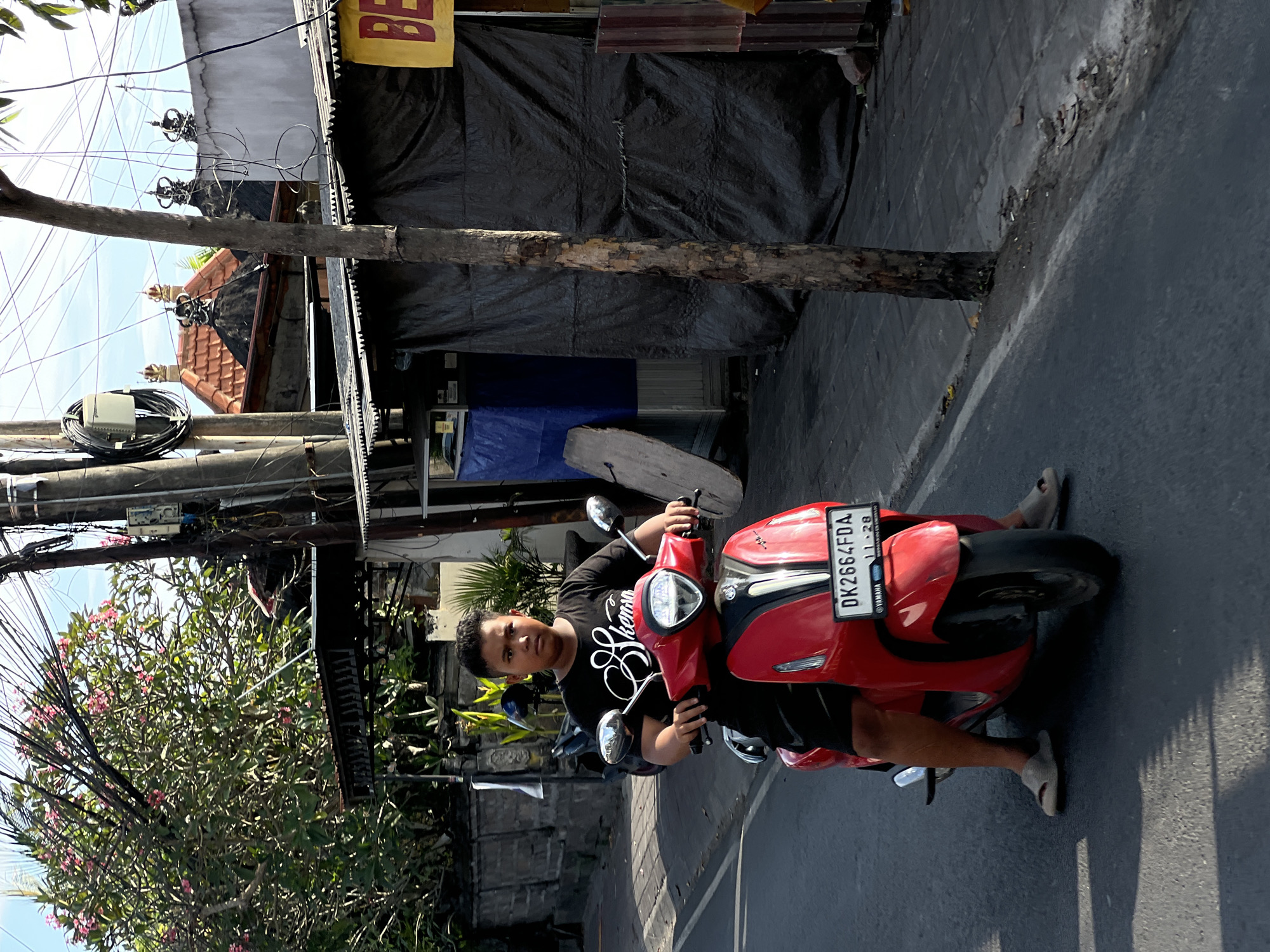A young boy rides a red scooter down a paved street in North Kuta, Indonesia. He is wearing a black t-shirt with white lettering and black shorts, along with light-colored sandals. The scooter has a license plate that reads "DK 2664 FDA". The street is lined with buildings, some covered in dark tarps, and overhead power lines are visible. A tree trunk stands on the right side of the frame. The sun casts shadows on the road.
