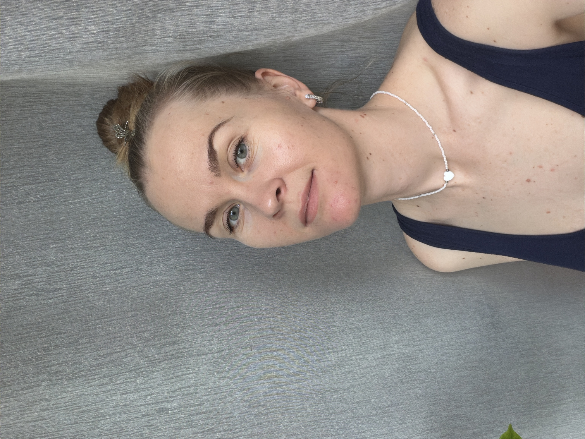 A fair-skinned woman with blonde hair pulled into a messy bun with a silver clip is shown from the chest up, leaning her head against a textured grey backdrop. She is wearing a dark blue sleeveless top and a white beaded necklace with a white heart pendant. She has blue-green eyes, fair skin with freckles and some redness on her cheeks and chin, and light brown eyebrows. A silver hoop earring with small stones is visible on her right ear. The lighting is even and appears to be natural daylight. The background is a plain, vertically textured grey curtain or fabric. There is no discernible information in the image that would indicate a specific location in Khabarovsk, Russia.