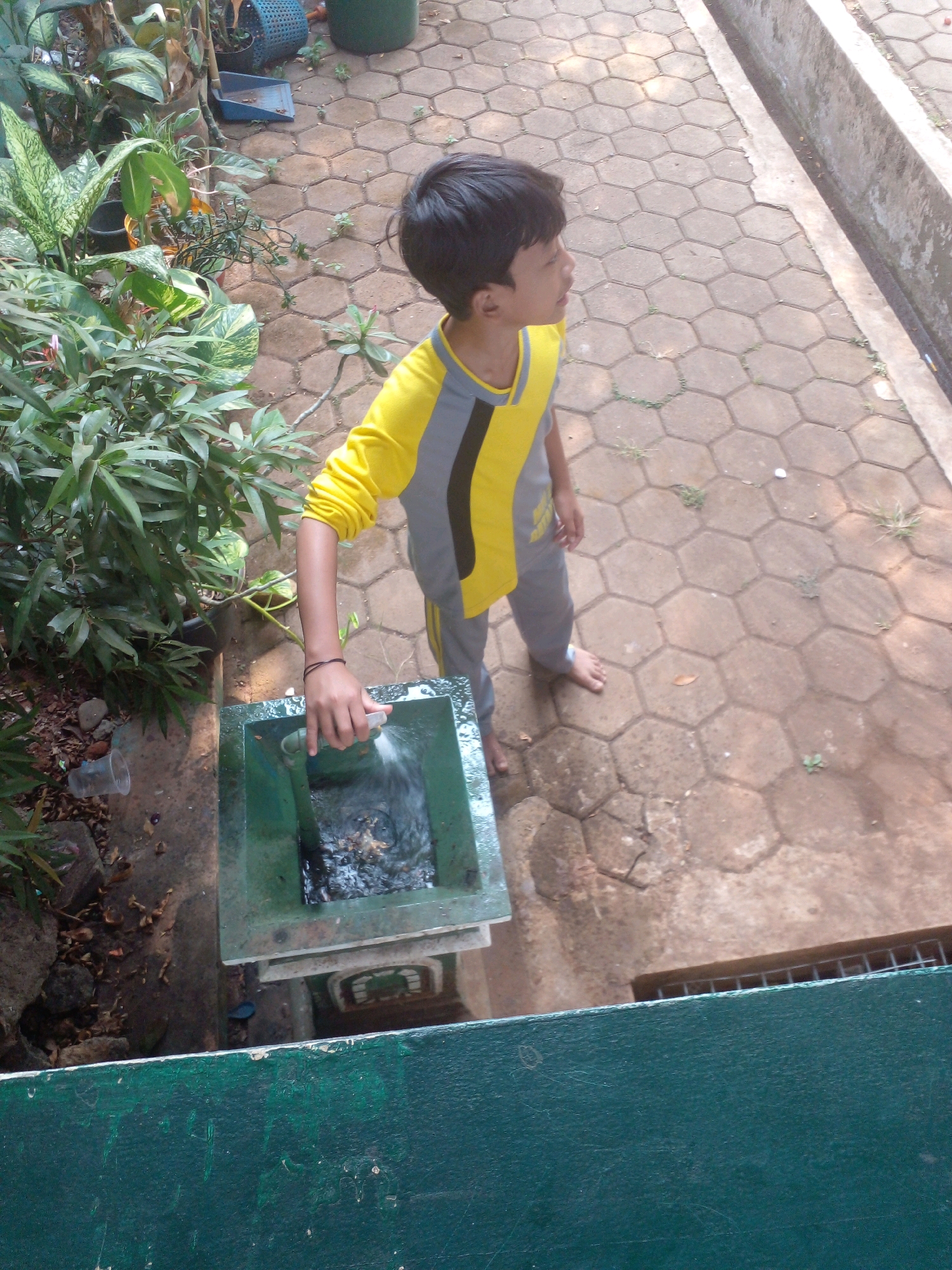 An outdoor scene shows a young boy standing barefoot on a hexagonal-tiled pathway in Banyuputih, Indonesia. He is wearing a yellow and grey long-sleeved shirt and grey pants. In his right hand, he holds a white spray bottle, from which he is spraying water into a green, rectangular water basin. The basin has a pipe and is filled with murky water. To the boy's left, lush green plants are visible, with some dry leaves scattered on the ground. The pathway is bordered by a concrete structure on the right side.