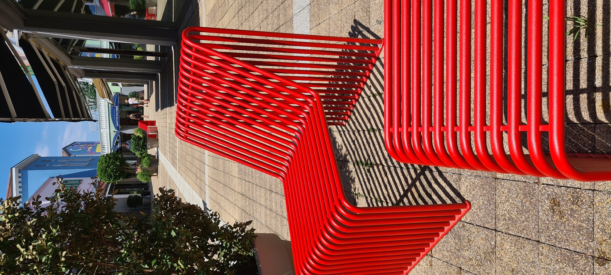 This image depicts a modern, bright red, tubular metal bench placed outdoors on a paved area. The bench has a distinctive zig-zag design, with multiple parallel tubes forming the seating surface and backrest. The scene appears to be a pedestrian area, possibly a shopping street or plaza, given the presence of shopfronts and awnings visible in the background. The architecture of the buildings suggests a contemporary design, with some featuring colorful facades.
In the background, several people can be seen walking, interacting, or browsing. One shop prominently displays the "Tommy Hilfiger" logo, providing a specific location cue. Lush green trees and potted plants are also visible, adding to the outdoor ambiance. The sunlight casts strong, striped shadows from the bench onto the pavement, indicating it is daytime, likely midday or early afternoon due to the sharp shadows. The sky is clear and blue, suggesting fair weather.
The overall impression is one of a pleasant, sunny day in a well-maintained urban or commercial outdoor space, likely in Aiello del Friuli / Dael, Italy, as indicated by the location context. The vibrant red of the bench contrasts with the neutral tones of the paving and the natural greens of the foliage, making it a focal point of the composition.