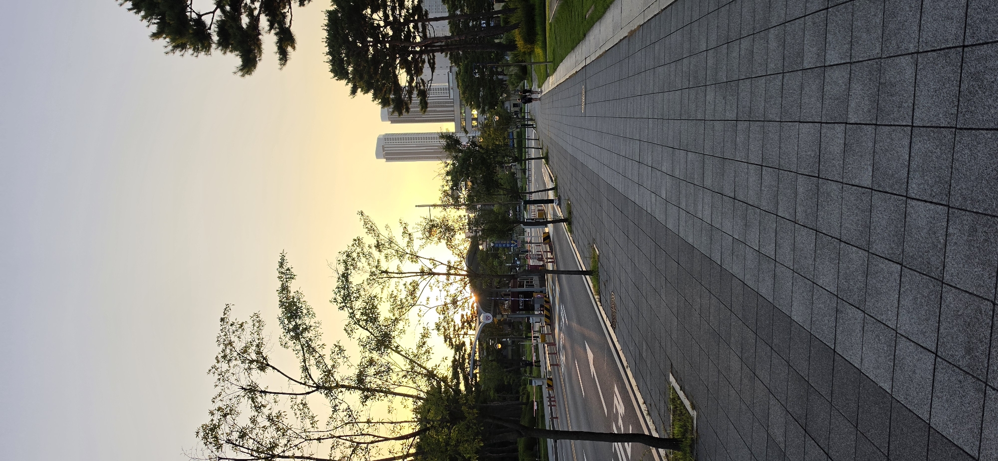 The image shows a pedestrian walkway beside a road in Cheongju-si, South Korea, during a sunset. The sky is a soft yellow and orange, indicating the sun is low on the horizon. On the left, a paved road with lane markings is visible, with trees and greenery lining the side. A toll booth structure is present further down the road. On the right, the wide pedestrian walkway is made of square gray tiles, with a grassy area beyond it. Several tall trees, including pine trees, are scattered throughout the scene. In the distance, modern high-rise buildings are silhouetted against the sky. Two people are walking away from the camera on the pedestrian walkway. The overall atmosphere is serene, capturing the end of the day.