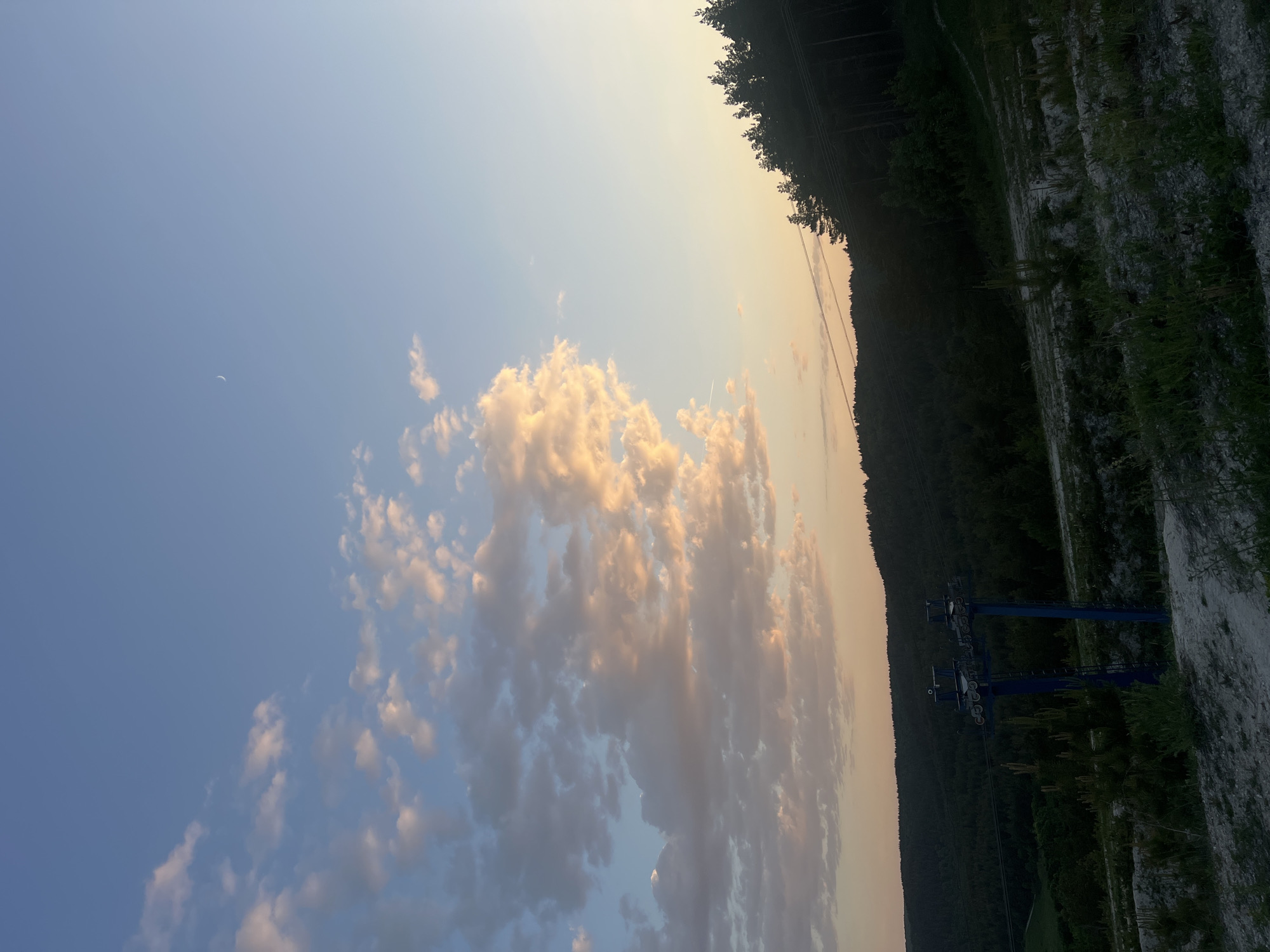 The image shows a landscape in Khvalynsk, Russia. In the sky, cumulus clouds illuminated by the setting sun, with a thin crescent moon visible. The right side of the image features a dark forested hillside with a chairlift structure. A dirt and gravel area is in the foreground. The overall lighting suggests either sunset or sunrise.