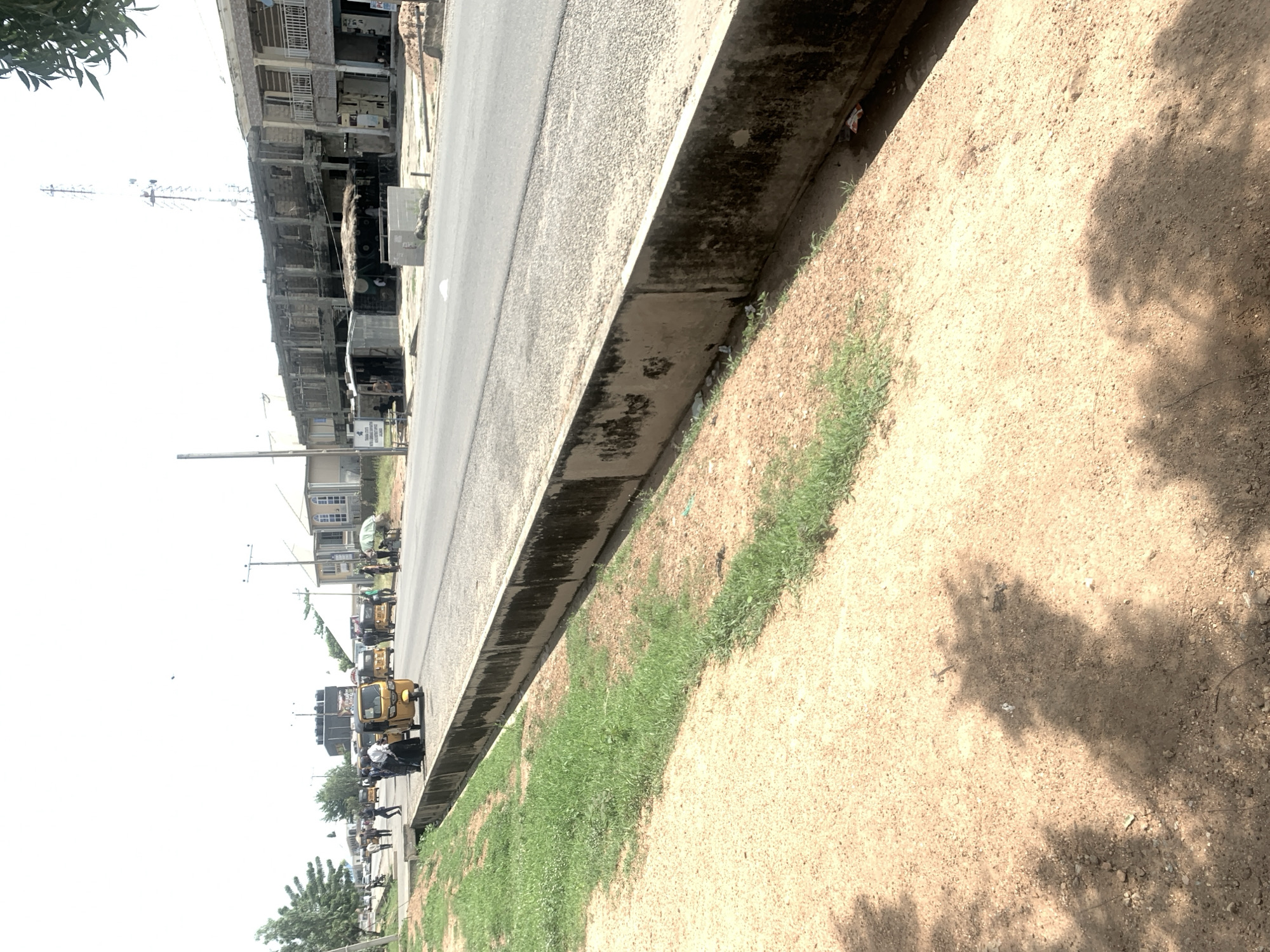 The image depicts a street scene in Jalingo, Nigeria. A paved road runs alongside a raised concrete barrier, with a sandy embankment and patches of grass on the right. The scene is dominated by several yellow tricycles, commonly known as "keke napeps," parked and in motion along the roadside. People are visible interacting with the tricycles and walking along the street. In the background, there are multi-story buildings, some appearing to be shops or residential apartments, lining the road. A telecommunications tower is also visible in the distance. The sky is bright, suggesting daytime, and the weather appears to be clear. The overall impression is that of a bustling urban environment with typical modes of transportation.