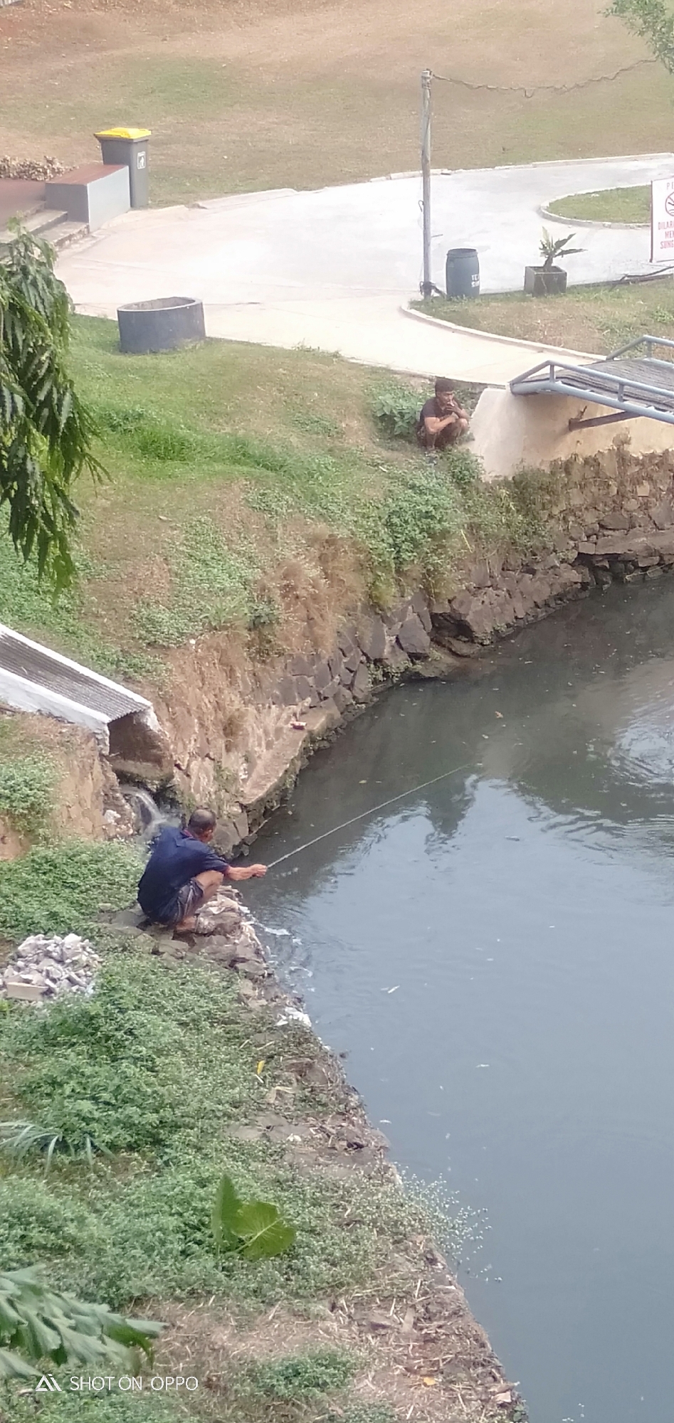 In Depok, Indonesia, two men are observed by a canal. One man, wearing a dark blue shirt and patterned shorts, is squatting on the rocky bank of the canal, actively fishing with a line extending into the murky water. Adjacent to him, further up the grassy embankment, another man is also squatting, appearing to be resting or observing. The canal bank is lined with rough stones, and the water itself appears somewhat polluted. Overgrown vegetation covers parts of the embankment. In the background, a paved pathway, a trash receptacle, and a cylindrical concrete structure are visible on a higher grassy area. A metal bridge or walkway spans across the canal in the middle ground.