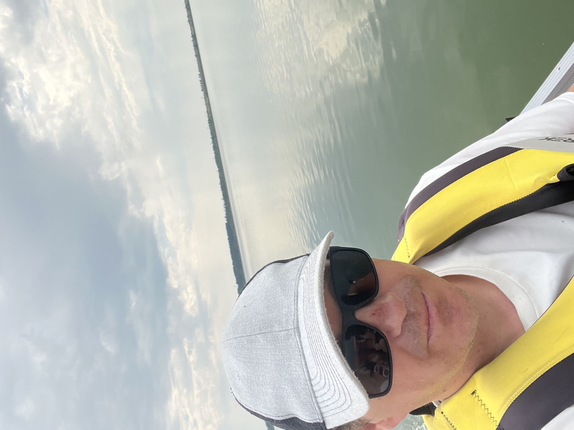 A selfie taken from a low angle shows a man wearing a grey baseball cap with a black mesh back, black sunglasses, and a yellow and black life vest over a white t-shirt. He is on a body of water, possibly a lake, with a shoreline visible in the distance. The sky is overcast with clouds. The water is calm with reflections of the sky. The location is identified as Beregovoye, Kazakhstan.
