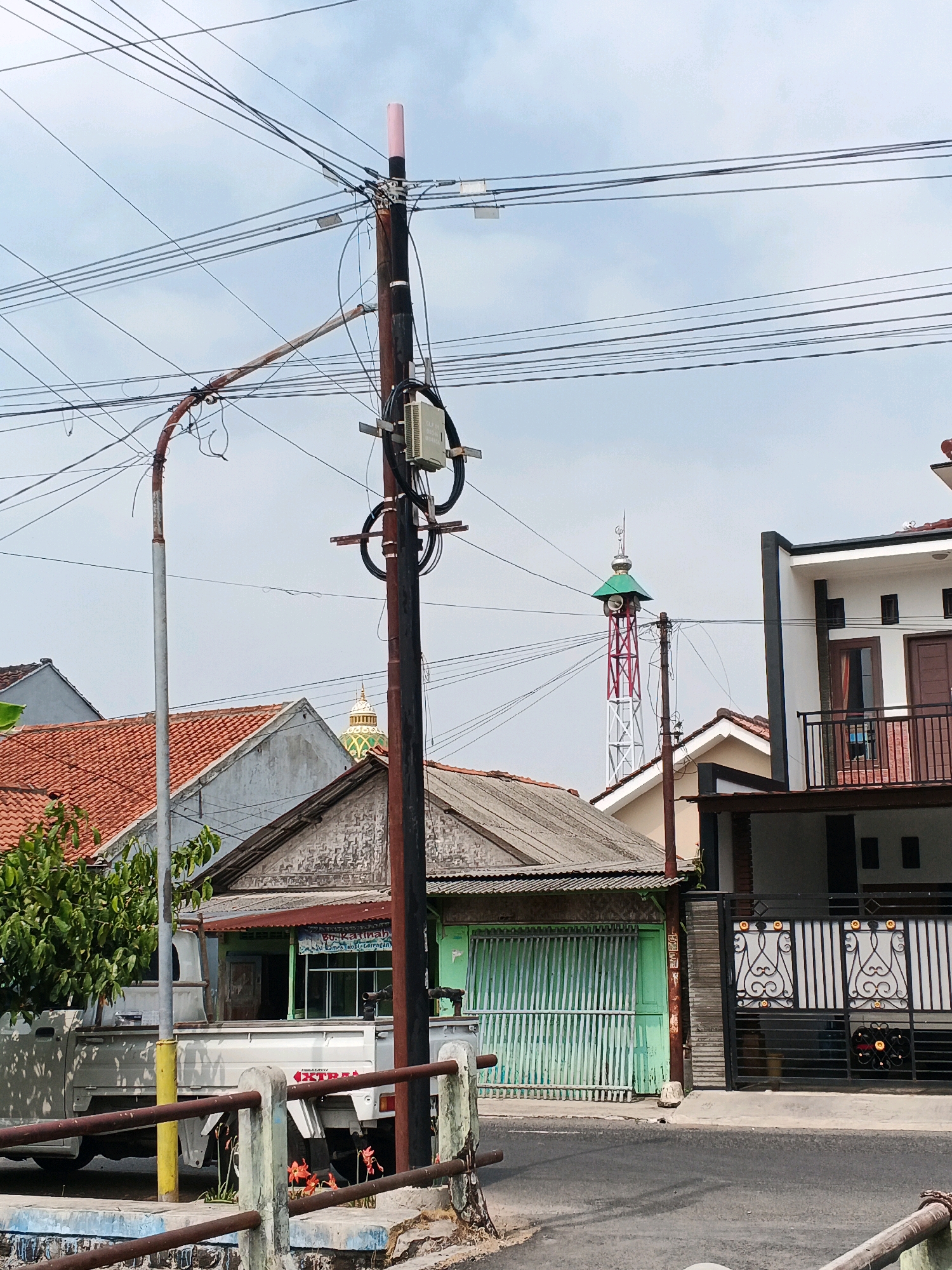 The image depicts a street scene in Kelurahan Tegalkamulyan, Cilacap, Indonesia. In the foreground, a damaged metal fence runs horizontally across the bottom of the frame, with a yellow pole on the left and a weathered brown pole on the right. Behind the fence, a white pickup truck is parked on the left side of the street, partially visible.
Dominating the center of the image is a tall, dark brown utility pole supporting numerous electrical wires. A pink-capped antenna is attached to the top of the pole, and a white box-like equipment unit is mounted midway up. To the left of this pole, a separate, lighter-colored utility pole with a curved, rusted arm extends across the frame.
In the background, several buildings are visible. On the far left, a building with an orange-tiled roof is seen. Adjacent to it is a building with a light gray, corrugated roof and green shutters. Further back, a distinctive red and white lattice tower with a green-roofed structure at its apex stands tall, resembling a minaret or communication tower, with two speakers mounted on its sides. To the right of the tower is a two-story building with white walls, dark trim, and a black metal gate. The sky is overcast with scattered clouds. The overall scene suggests a typical urban or suburban environment with active infrastructure.