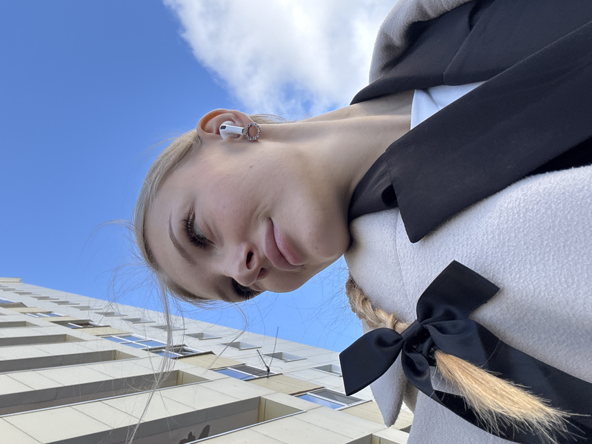 A young woman with blonde hair and makeup is taking a selfie outdoors. She is looking up and to the right, with a slight smile. She is wearing a light-colored coat over a dark jacket and a white top. A black ribbon is tied around her braided blonde hair, which extends out from under her coat. She has a white earbud in her right ear and a circular earring. In the background, a modern building with many windows rises up towards a blue sky with scattered clouds. This scene is located in the city of Perm, Russia.