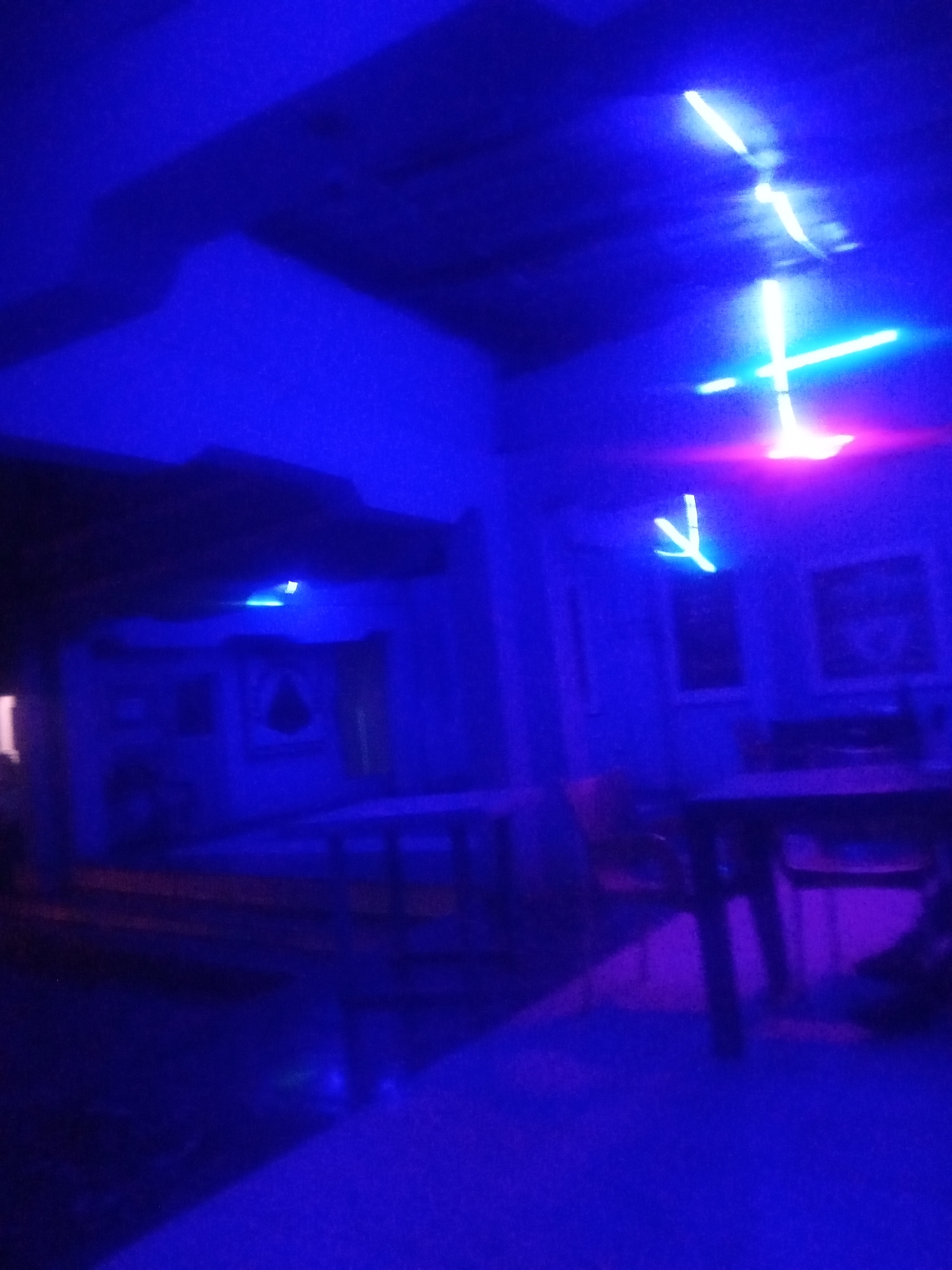 The image depicts an interior scene illuminated by vibrant blue and pink neon lights. The lighting creates strong shadows and washes out details, giving the environment a somewhat abstract and moody feel. In the upper portion of the image, several bright, geometric light fixtures are visible, possibly neon tubes arranged in linear and cross shapes.
In the mid-ground, there appears to be furniture, including what looks like tables and chairs, though their forms are indistinct due to the low light and color saturation. Some framed pictures or artwork are discernible on a wall in the background. The overall impression is that of a dimly lit entertainment venue or a decorated space with a contemporary lighting design. The specific location in Sabon Gari, Nassarawa, Nigeria, is not visually evident from the image itself, but the description is of the scene within that context.