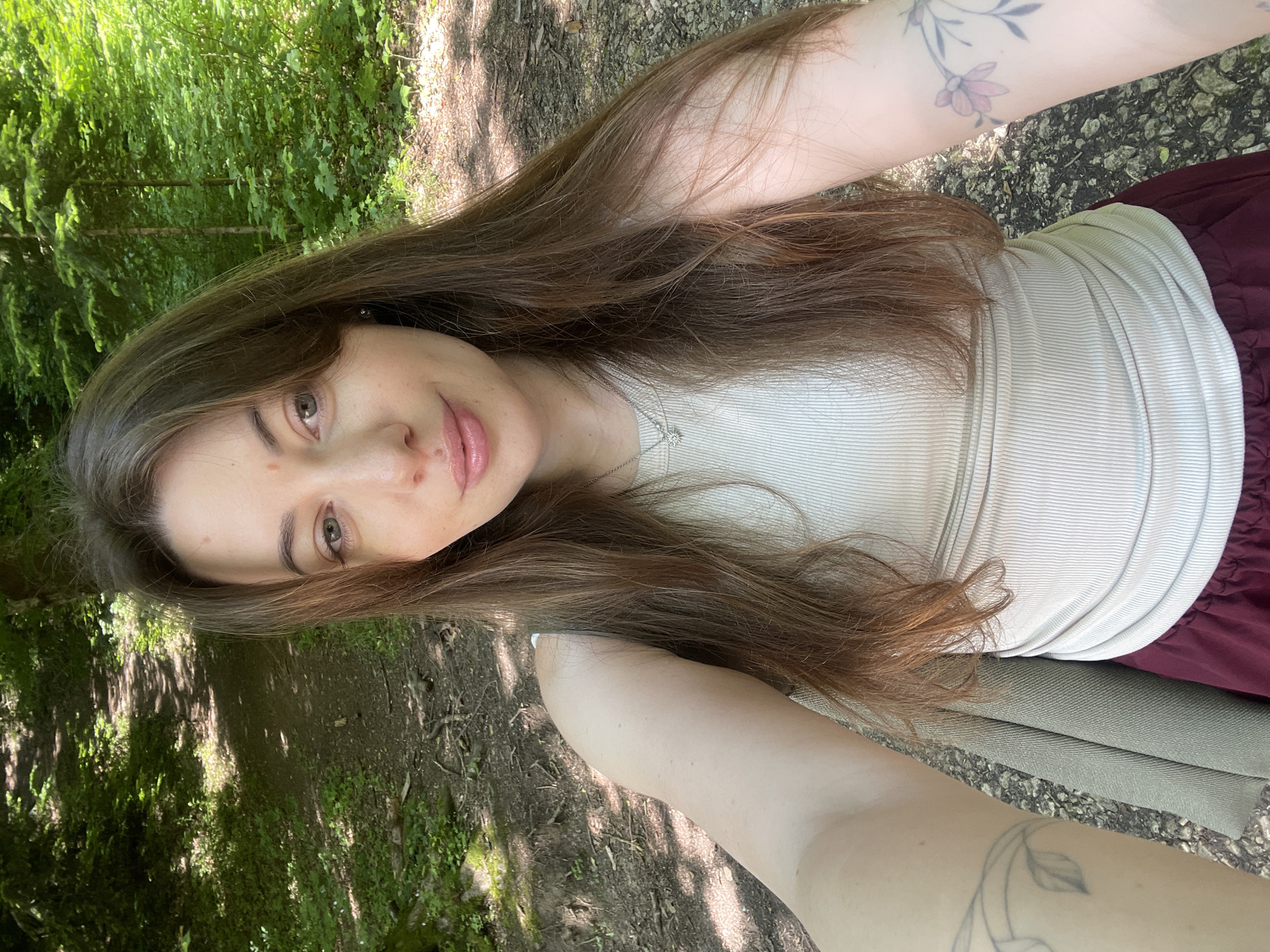 In Guzeripl', Russia, a young woman with long brown hair, wearing a beige ribbed tank top and a maroon skirt, takes a selfie outdoors. She has a delicate necklace with a star pendant. Her right arm is visible, showing a tattoo of a flower. She is positioned in a forest clearing with a dirt path winding through it, shaded by lush green trees. Sunlight filters through the leaves, dappling the ground. The overall scene suggests a peaceful nature walk.