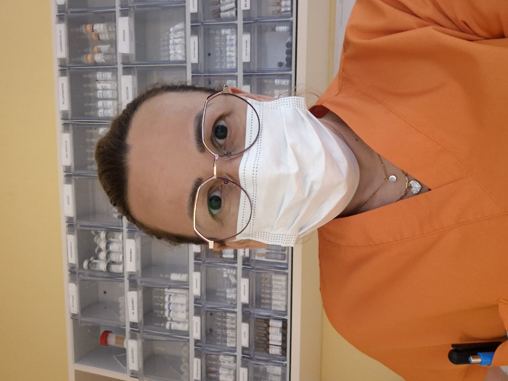 The image shows a medium close-up, selfie-style photograph of a person wearing an orange medical scrub top, a white face mask, and large-framed glasses. The person has their hair pulled back and is wearing two necklaces, one with a small circular pendant and another with a heart-shaped pendant. A blue and black pen is visible in the person's left breast pocket. Behind the person is a wall-mounted shelving unit filled with rows of clear plastic containers, each holding vials or ampoules of medication. The shelves have white labels with text that is partially visible but not legible. The background wall is painted a light yellow. The overall setting appears to be a medical or pharmacy environment. The lighting is bright and even.