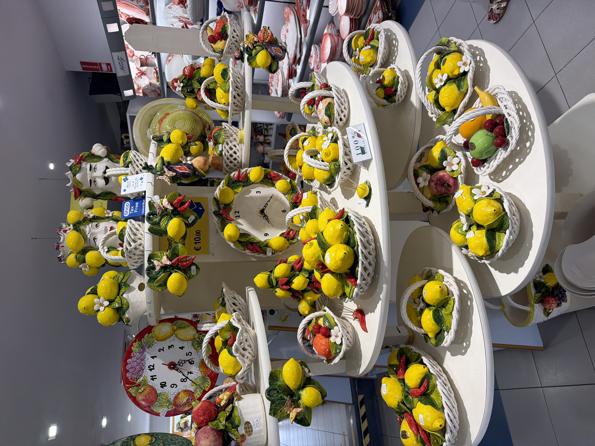 The image captures a shop interior in Ischia, Italy, showcasing a display of ceramic art, including clocks and wall-mounted baskets filled with vibrant, artificial lemons and other fruits. The items are arranged on multiple white tiered tables, suggesting a curated product selection. Clocks with lemon and fruit designs are visible on the shelves, while plates and decorative items are displayed further back. Prominent signage indicating "Tax-Free Shopping" and "100% Made in Italy" suggests the shop targets tourists.