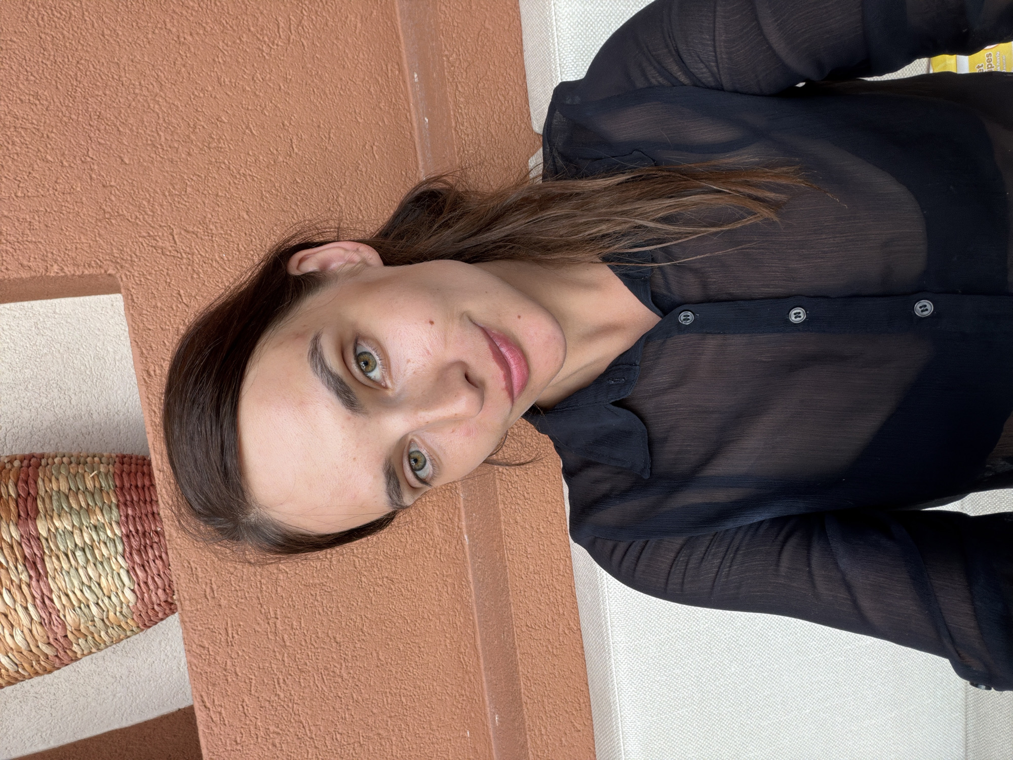 In Fethiye, Turkey, a young woman with brown hair and green eyes is captured in a selfie. She is wearing a sheer black blouse and a black bra visible underneath. The background features a textured terracotta wall and a woven basket. The woman is positioned on a white couch.