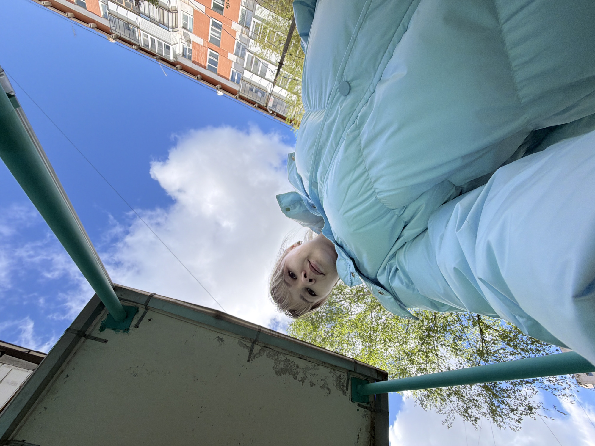 A person wearing a light blue puffer jacket is looking down at the camera. The camera is pointed upwards. Above the person is a building with an orange and white facade, partially obscured by tree branches. The sky is blue with scattered white clouds. Below the person is a drab, one-story structure with a green support beam extending outwards. The location may be Chaikovsky, Russia, based on prior knowledge.